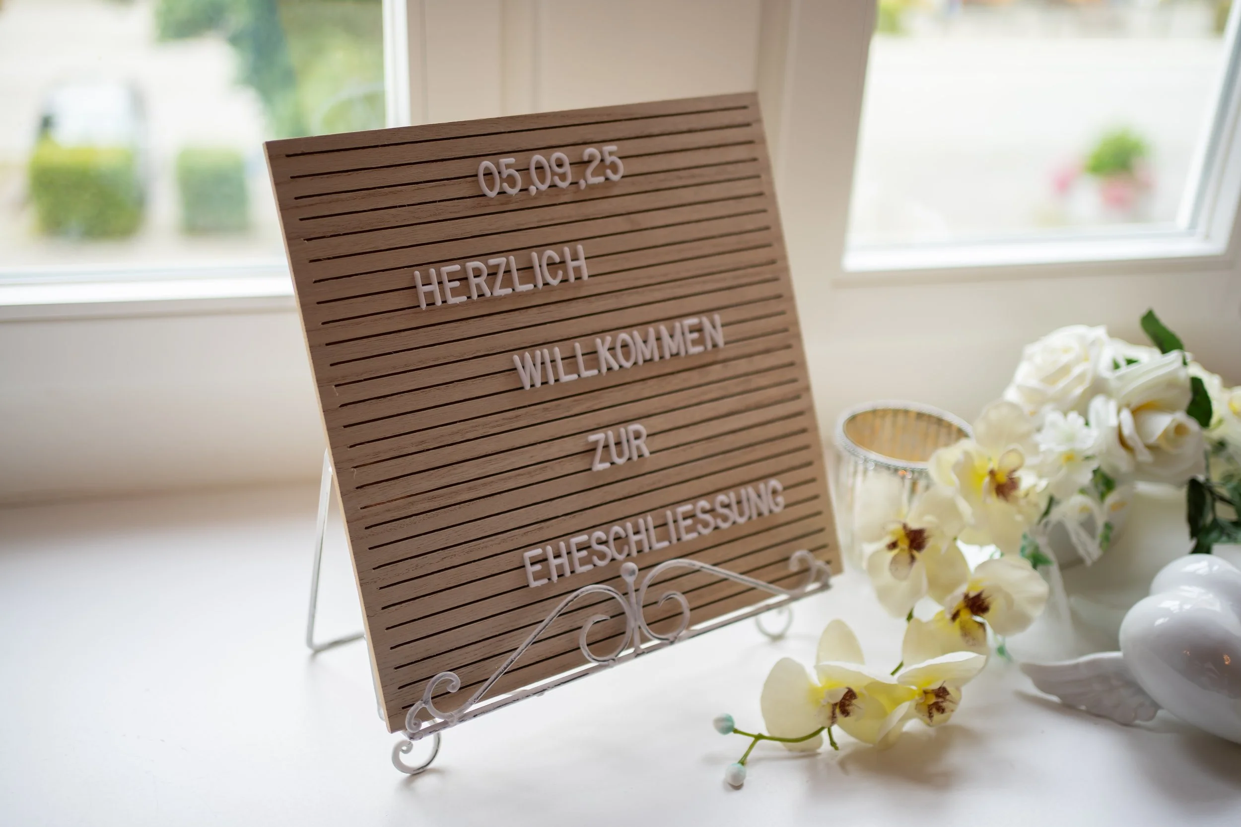 Ein Willkommensschild aus Holz mit weißen Buchstaben, das zur Hochzeitsfeier am 05.09.2025 einlädt, um die Gäste willkommen zu heißen, auf einem Tisch neben weißen Orchideenblumen und Kerzen, vor einem Fenster mit Blick nach draußen.