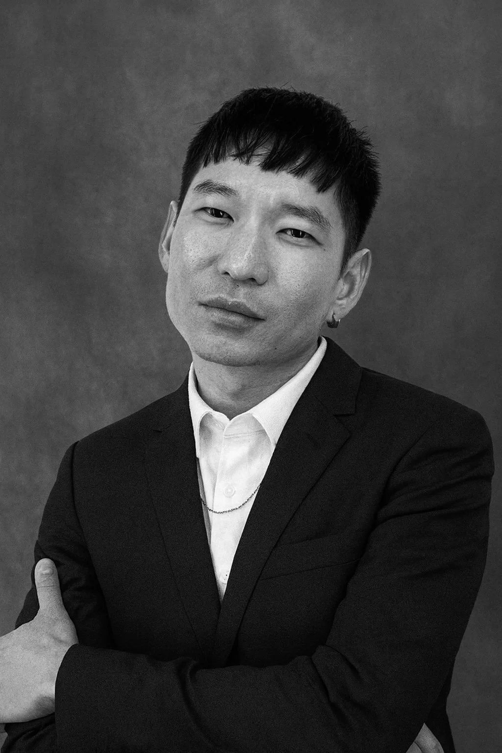 Black and white portrait of a young man wearing a suit jacket, white shirt, and chain necklace, with short, dark hair, crossing his arms and looking at the camera.