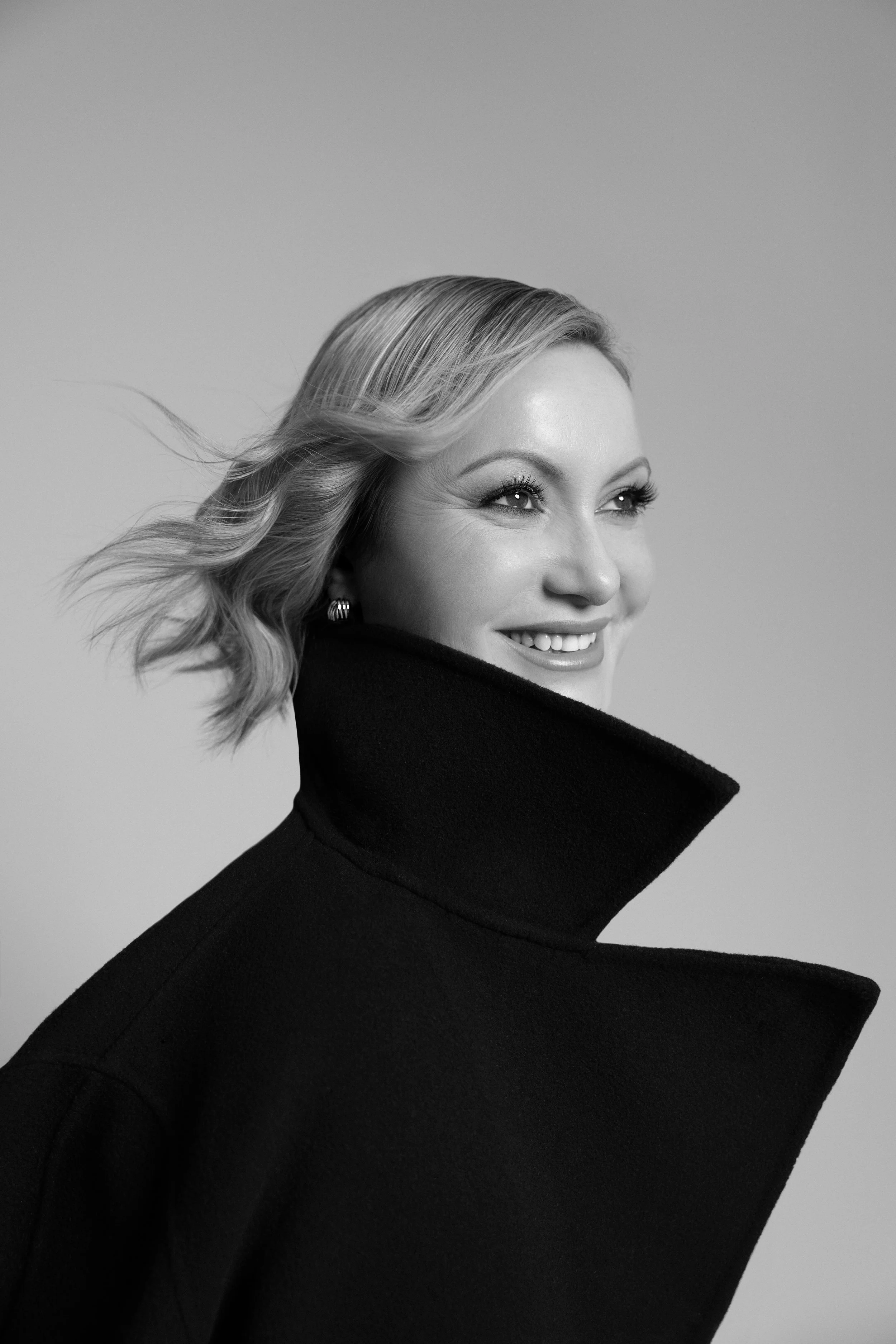 Black and white portrait of a smiling woman with styled blonde hair, wearing a high-collared coat.