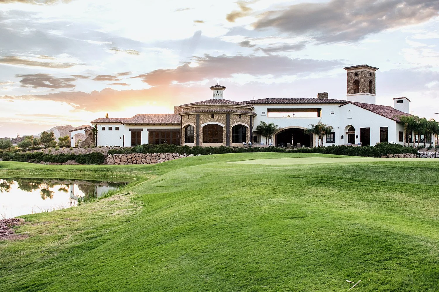 Luxury house with a manicured golf course in front and a small pond to the side at sunset with a cloudy sky.