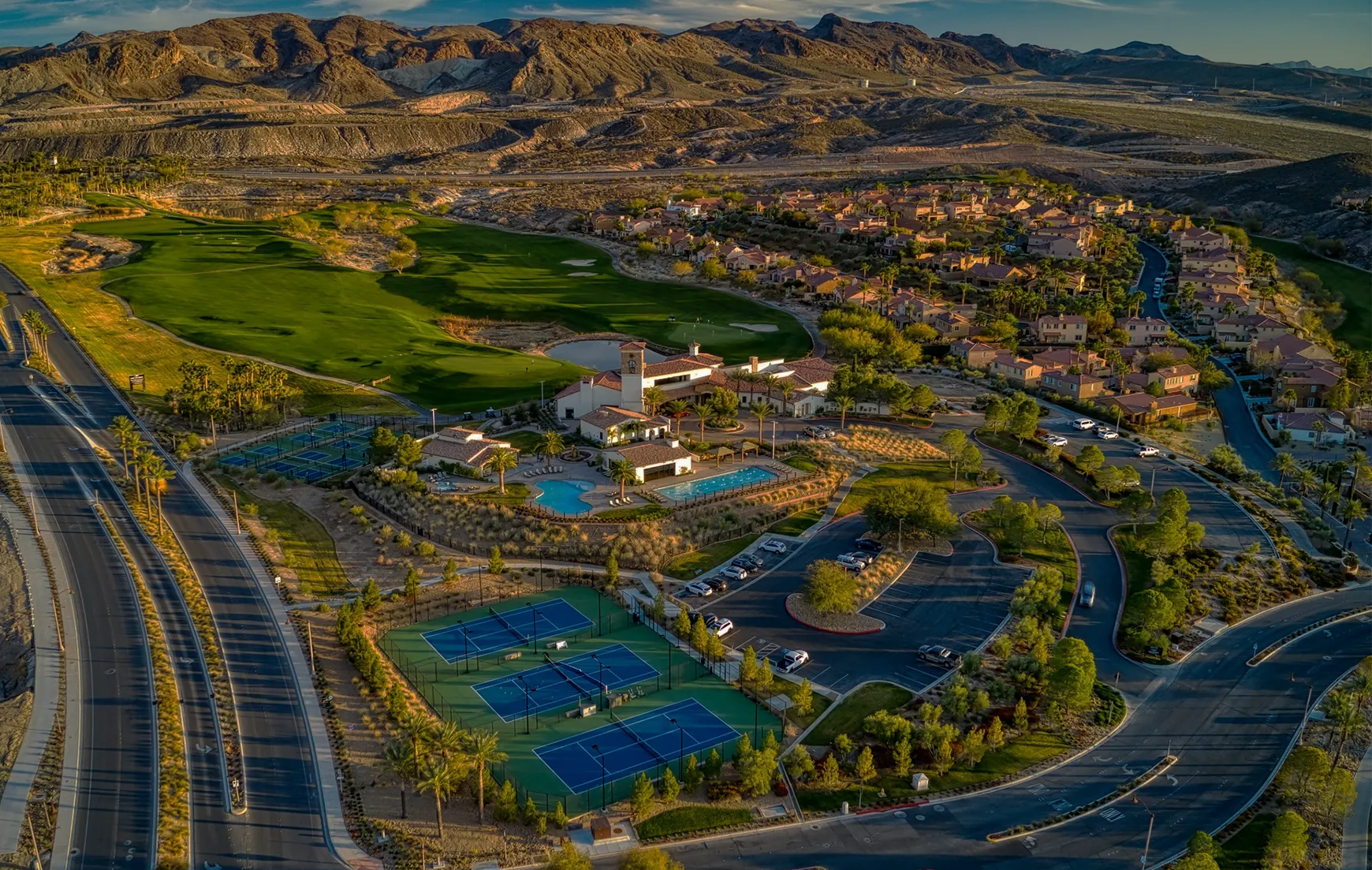 Aerial view of a residential community with a golf course, tennis courts, swimming pools, parking lots, and surrounding desert mountains at sunset.