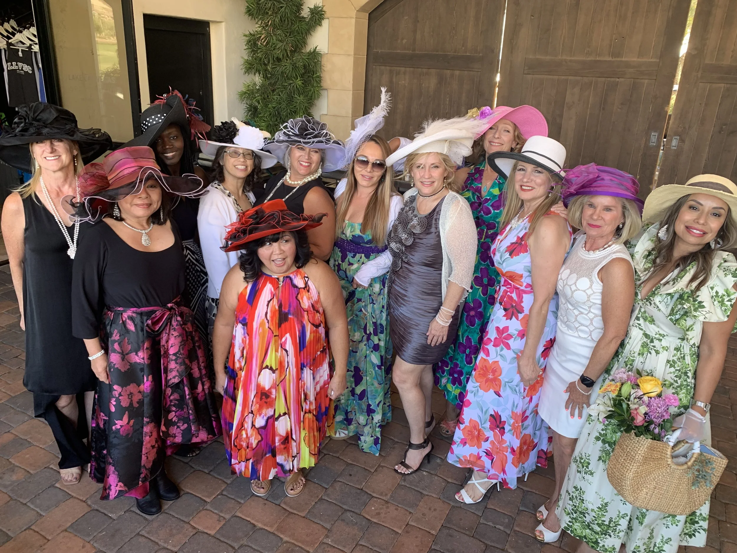 A group of women dressed in colorful and elegant dresses with large, elaborate hats, standing together outdoors on a brick patio.