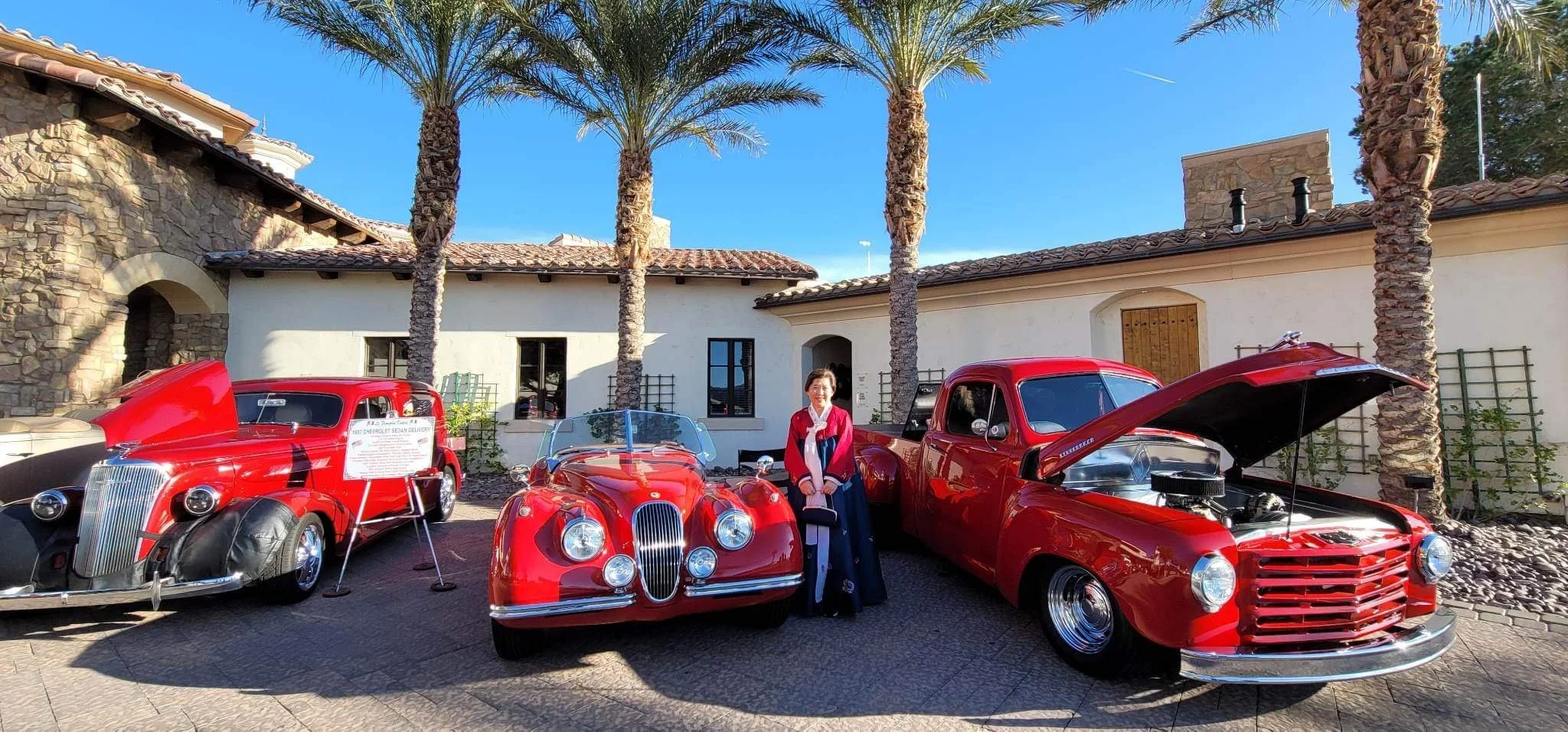 Lake Las Vegas Sport Club car show -  vintage red cars parked outdoors with a woman standing between the middle and right cars. The cars are displayed with their hoods open, and there are palm trees and a building with a tiled roof in the background 
