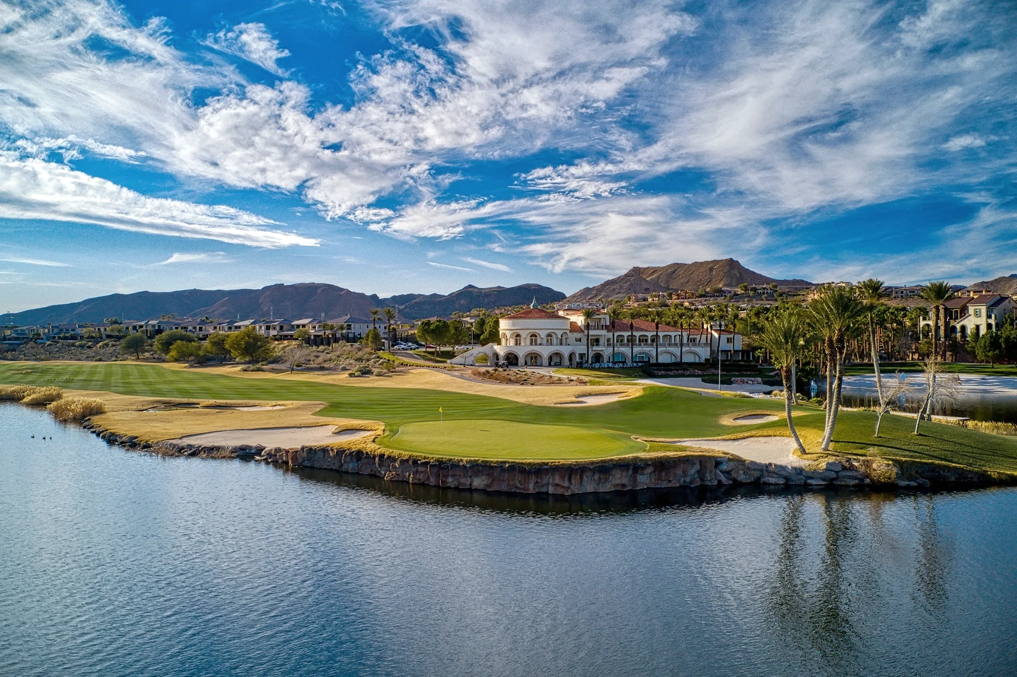 A scenic golf course with a water hazard and green, surrounded by palm trees and a large white resort building, set against a backdrop of mountains and a partly cloudy sky.