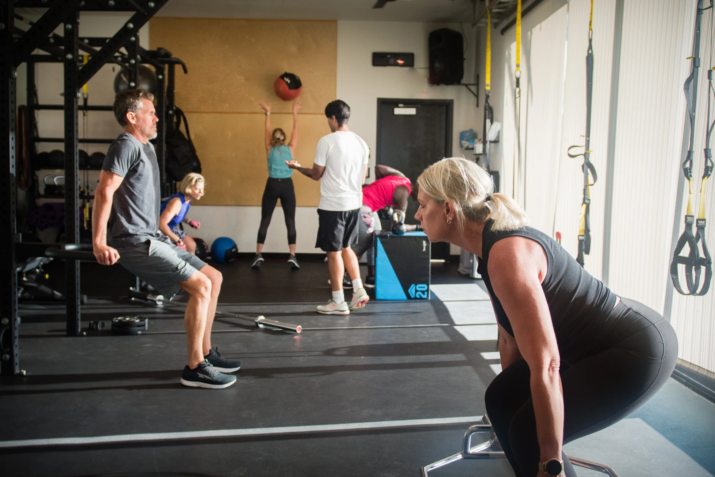 Lake Las Vegas Sport Club members working out at a gym, including a man doing squats, a woman in the foreground preparing to exercise, and others in the background engaged in different fitness activities.
