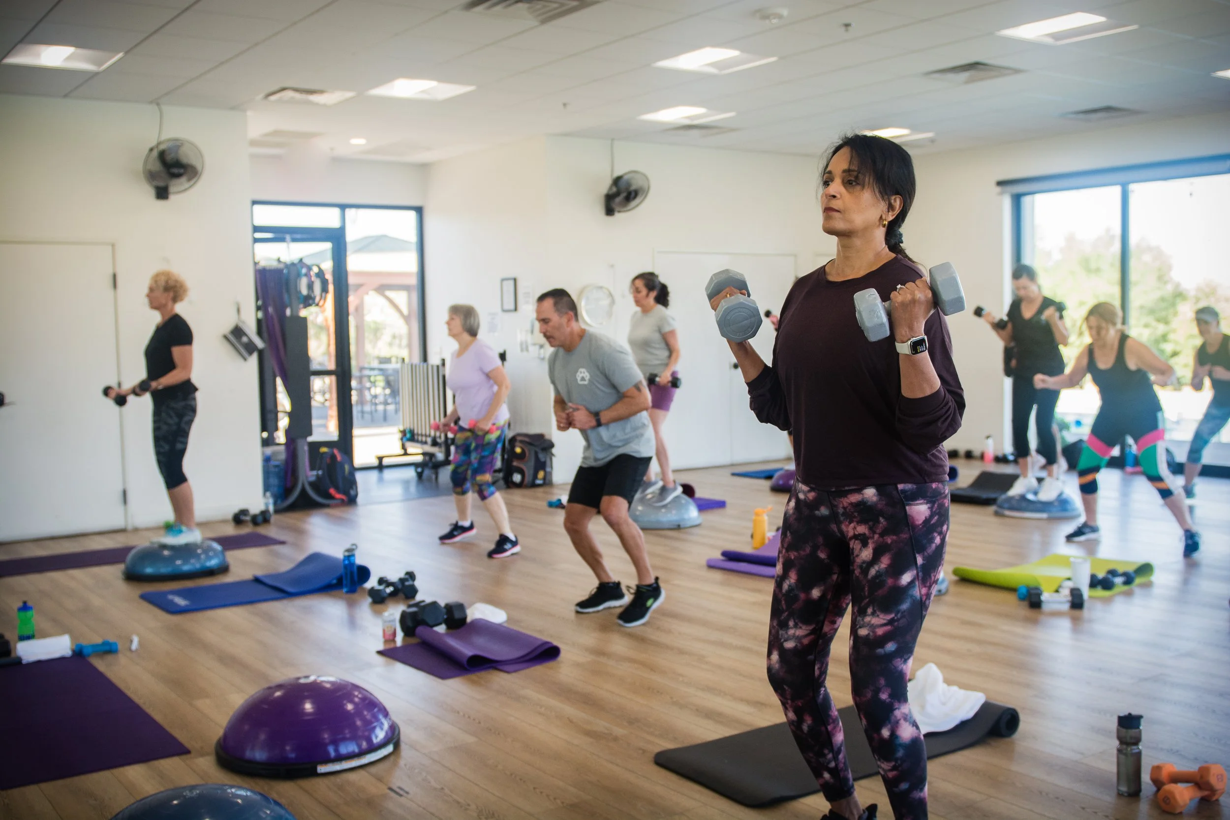 Lake Las Vegas Sport Club members working out in a fitness class, using dumbbells, balance balls, and mats in a spacious room.