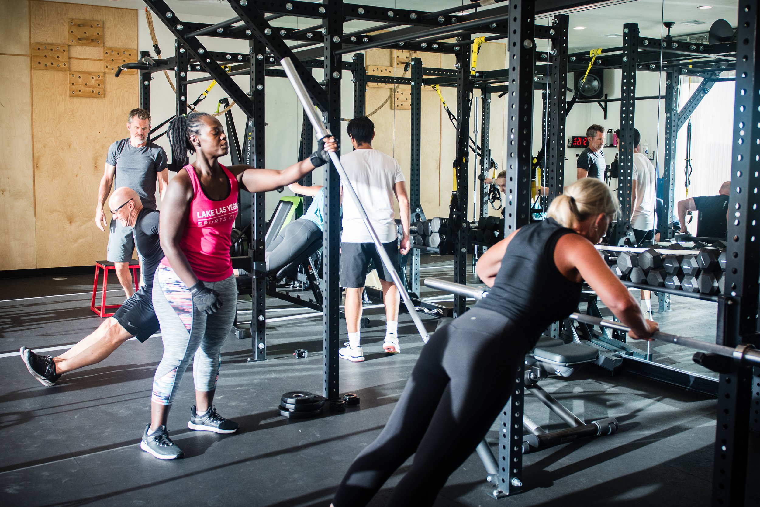 Lake Las Vegas Sport Club members exercising with gym equipment in a fitness center, including a woman doing a rowing exercise, a man doing a push-up, and others stretching or walking.