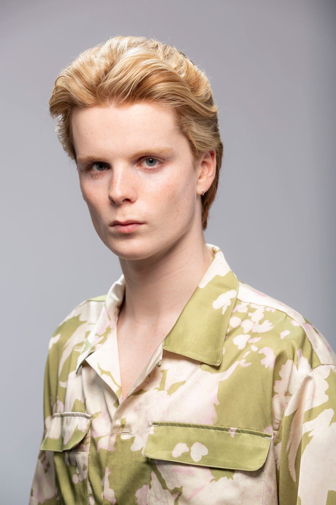 A young person with short, wavy, blonde hair, wearing a pastel-colored floral shirt, posing against a plain gray background.