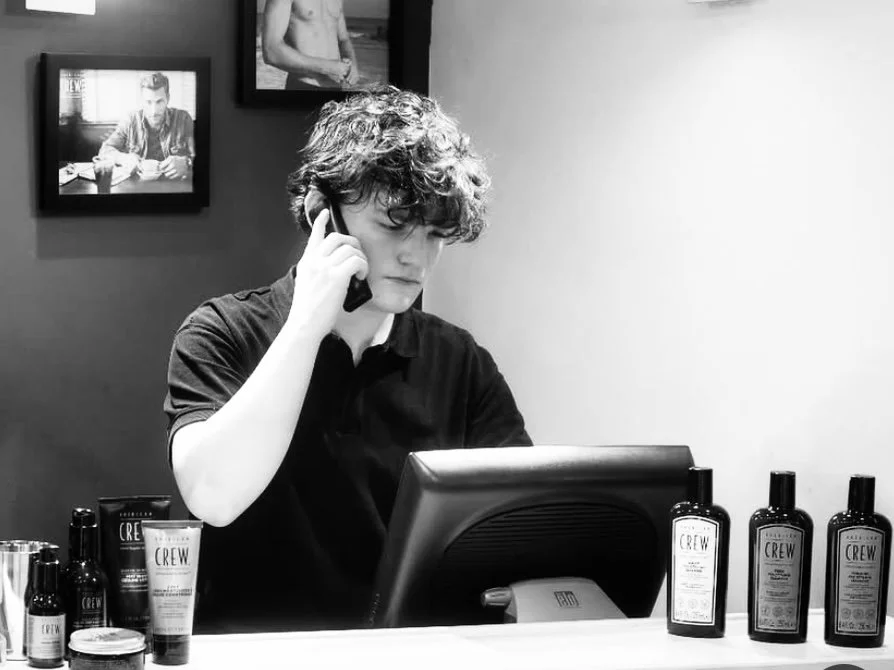 Young man with curly hair talking on a phone at a desk with various grooming products, in a black and white photo.