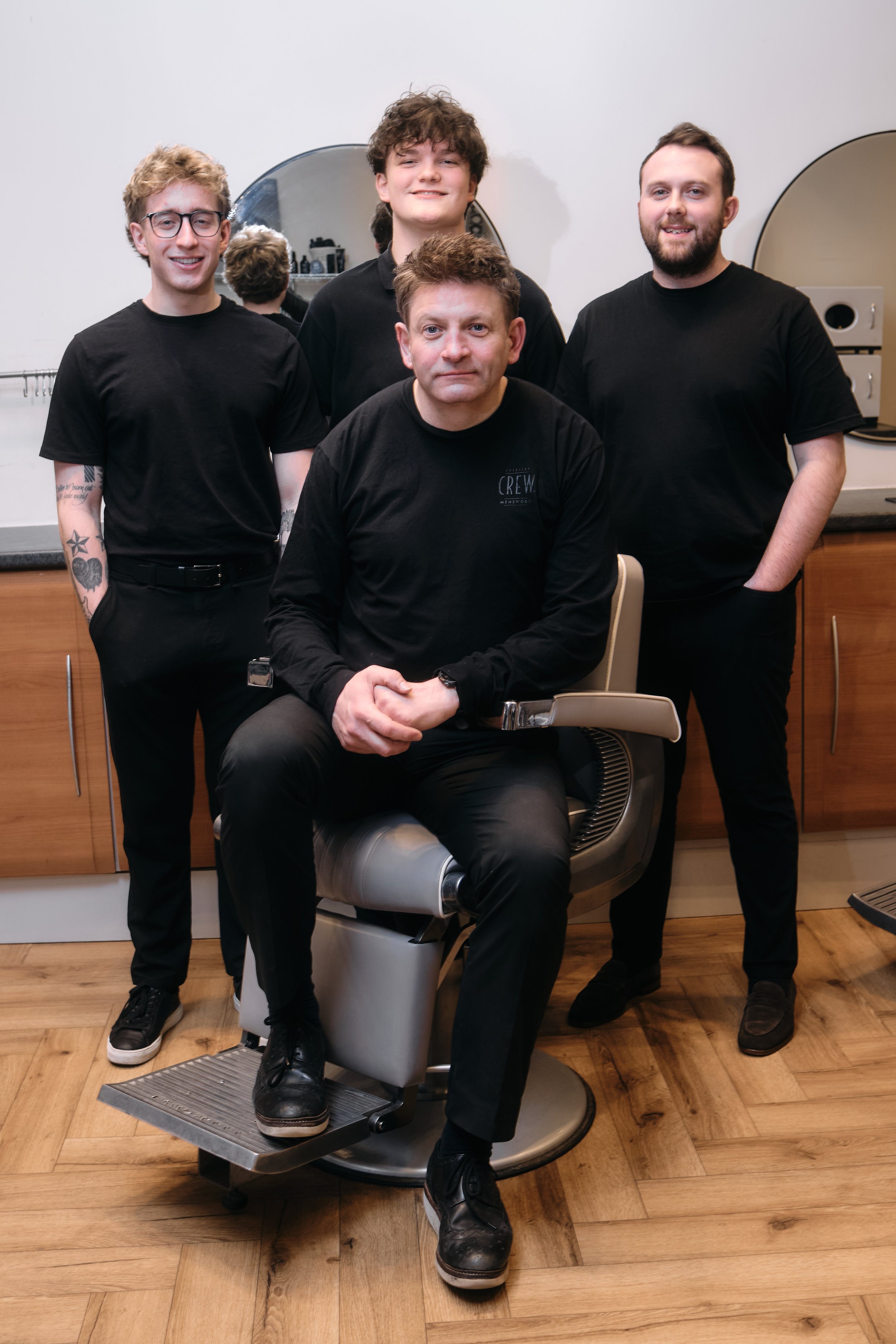 Four young men in black shirts posing in a barbershop, with one sitting in a barber chair and three standing behind him, smiling.