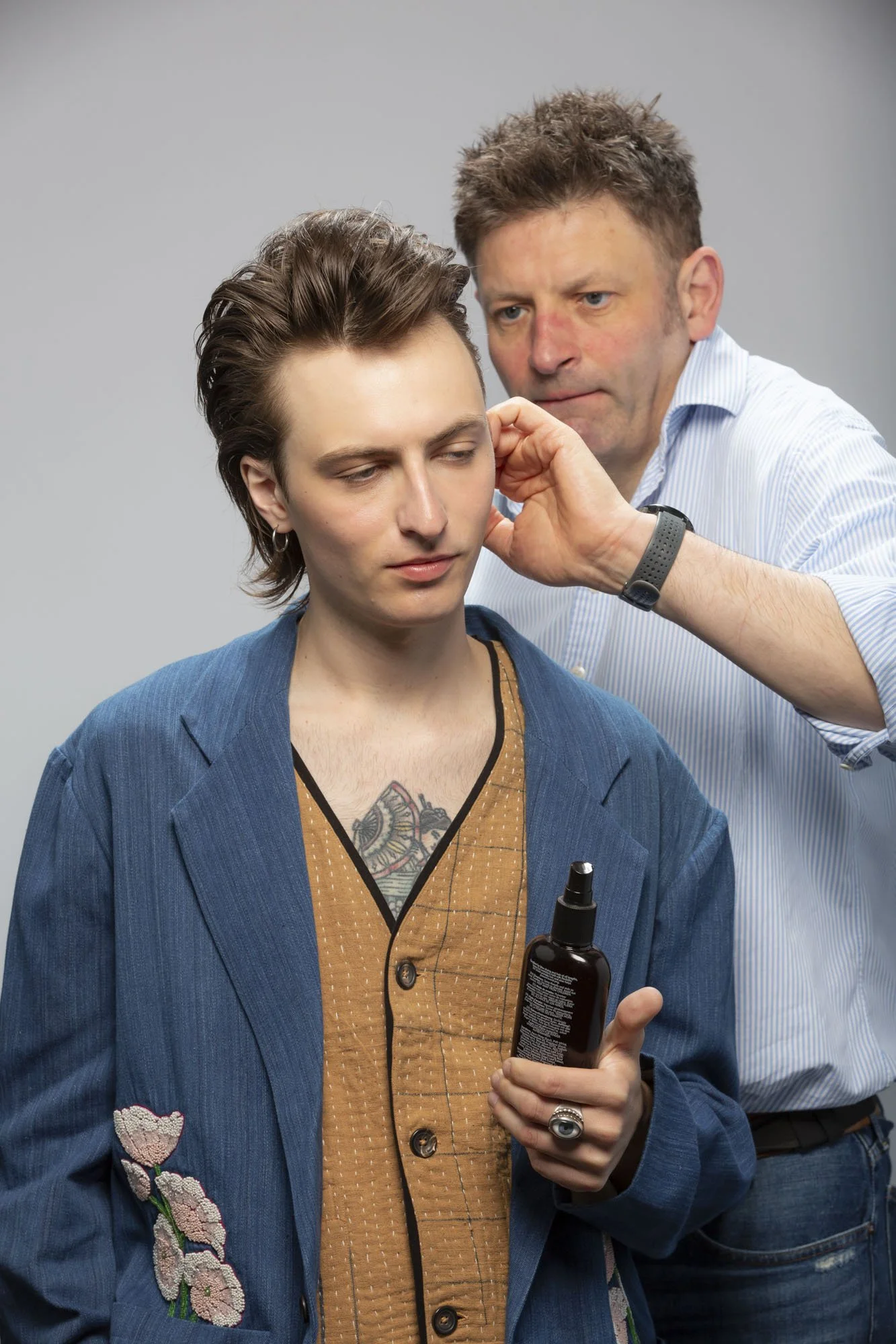 A man with short, tousled hair and a woman with short hair and earrings, in a studio. The man is adjusting the woman's ear while she holds a spray bottle, wearing a blue blazer with floral embroidery on the sleeve and a brown patterned shirt.