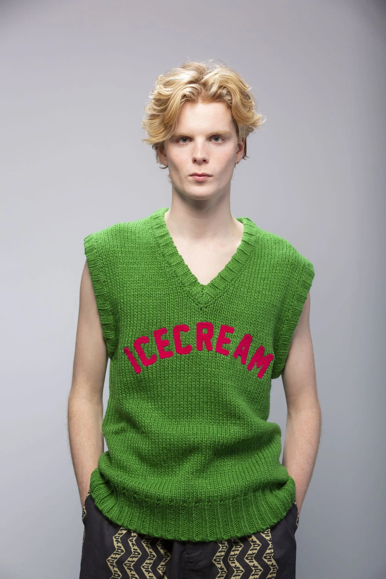 Person with short blonde curly hair wearing a green knitted sleeveless sweater with 'ICECREAM' written in pink across the front, and dark pants with a yellow pattern, standing against a plain gray background.