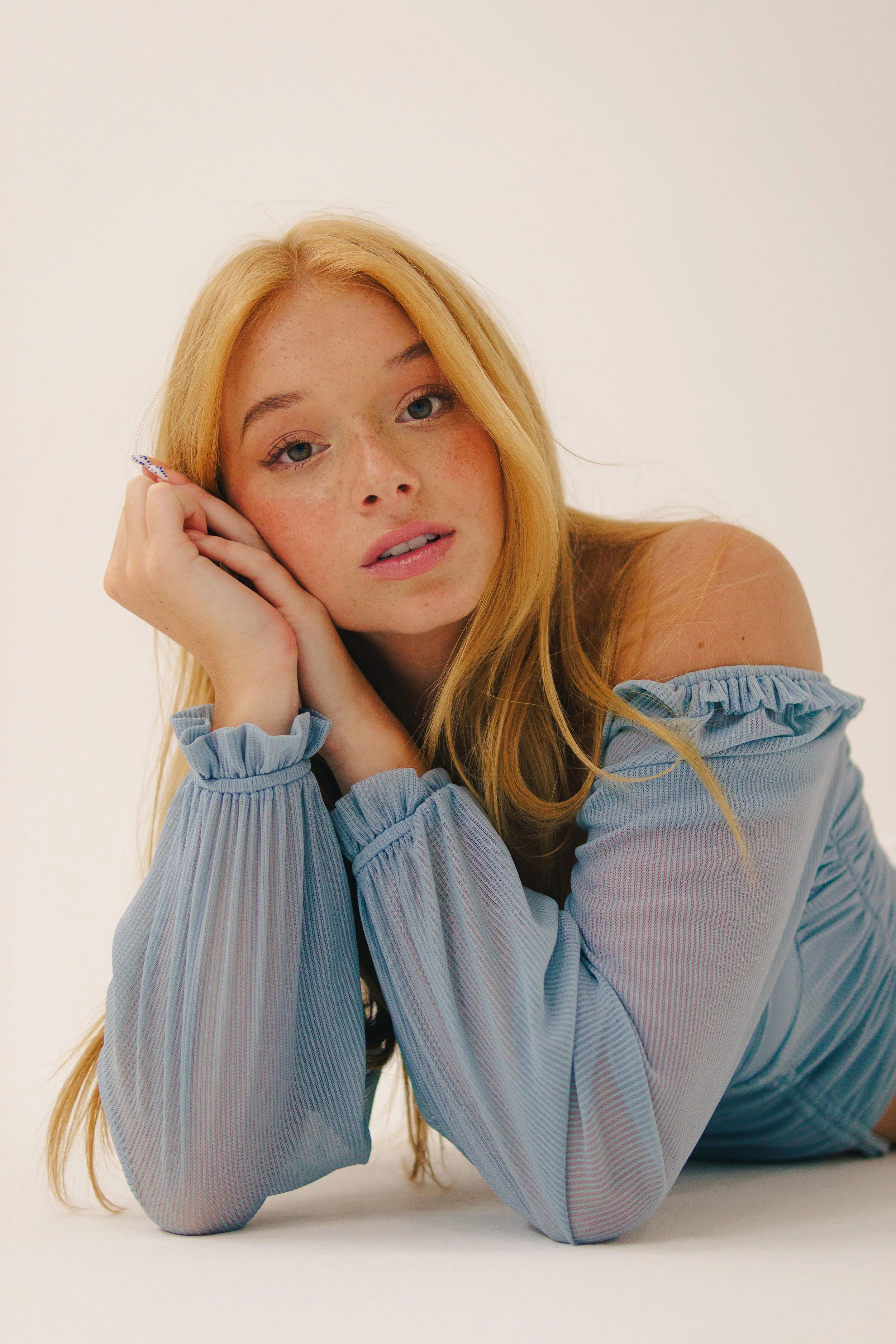 A young woman with long red hair and freckles lying on the floor, resting her head on her hand, wearing a light blue off-the-shoulder top, looking at the camera.