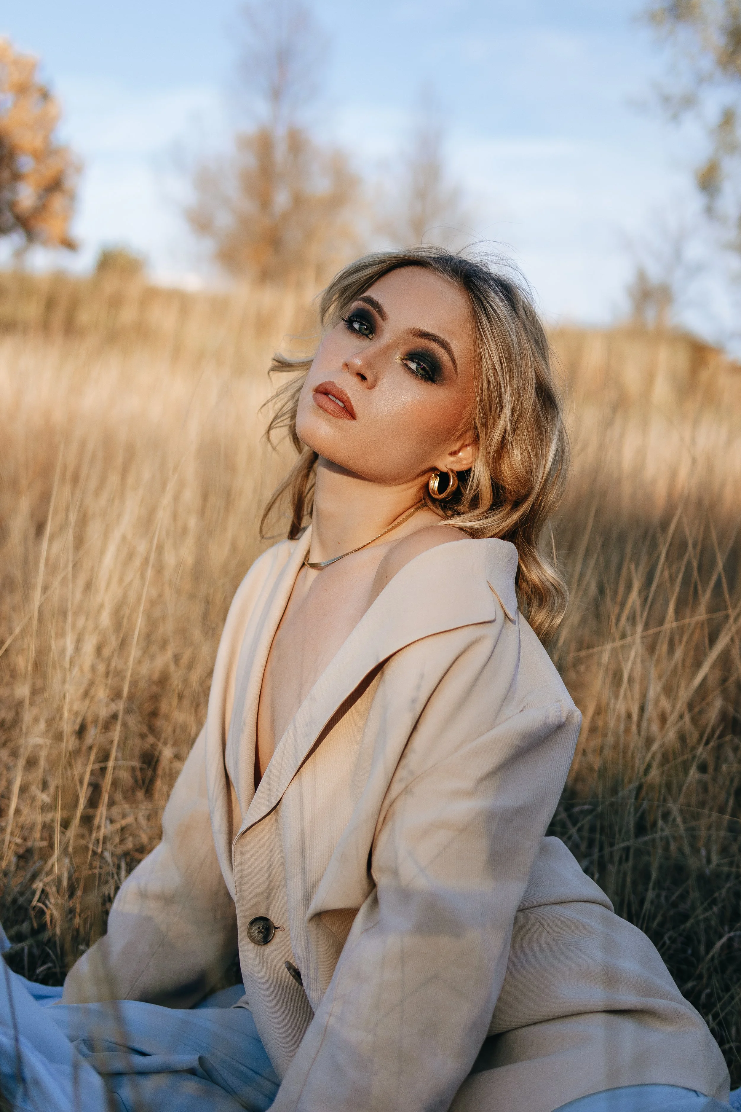 A woman with blonde hair and jewelry sitting in a field of tall, golden grass with a blue sky and trees in the background.