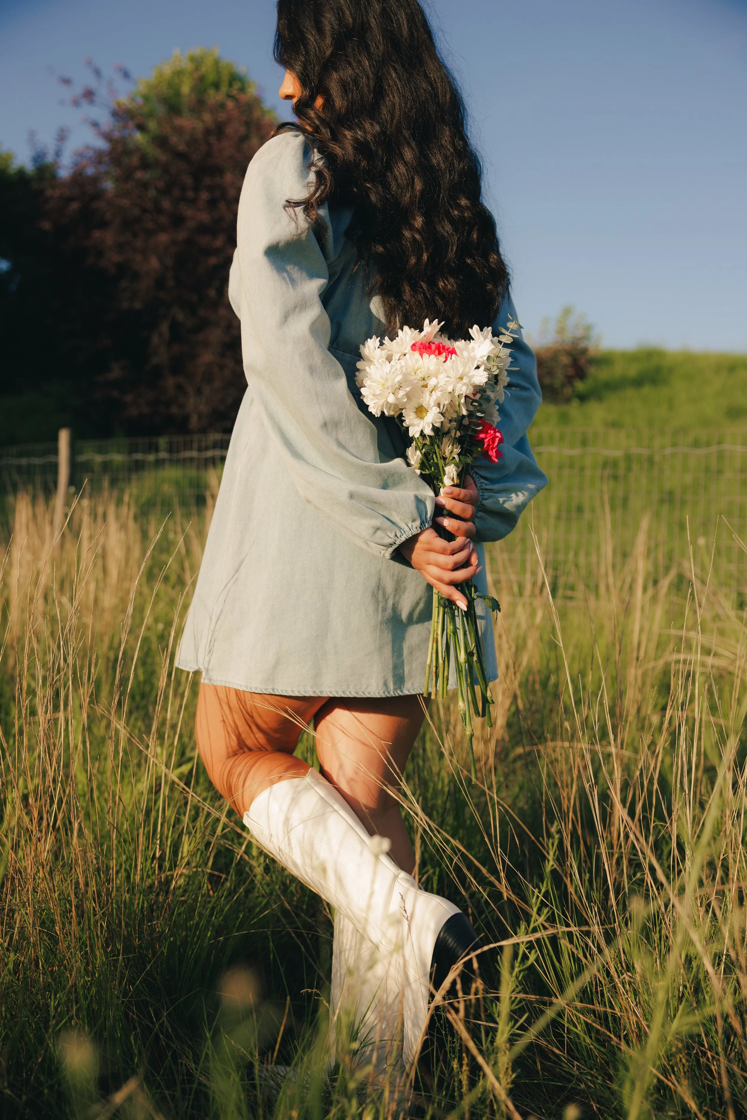 A woman with long dark hair stands in a grassy field, holding a bouquet of white and red flowers behind her back, wearing a long light-colored coat and white knee-high boots.