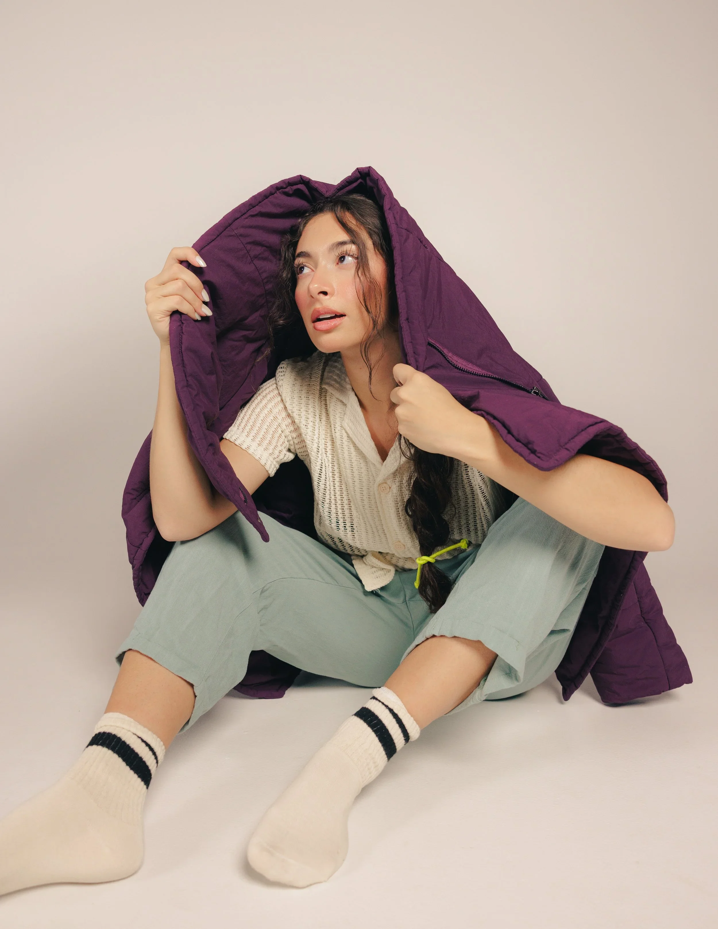 Young woman sitting on the floor with a purple jacket draped over her shoulders, wearing a beige knit shirt, light green pants, and white socks with black stripes.