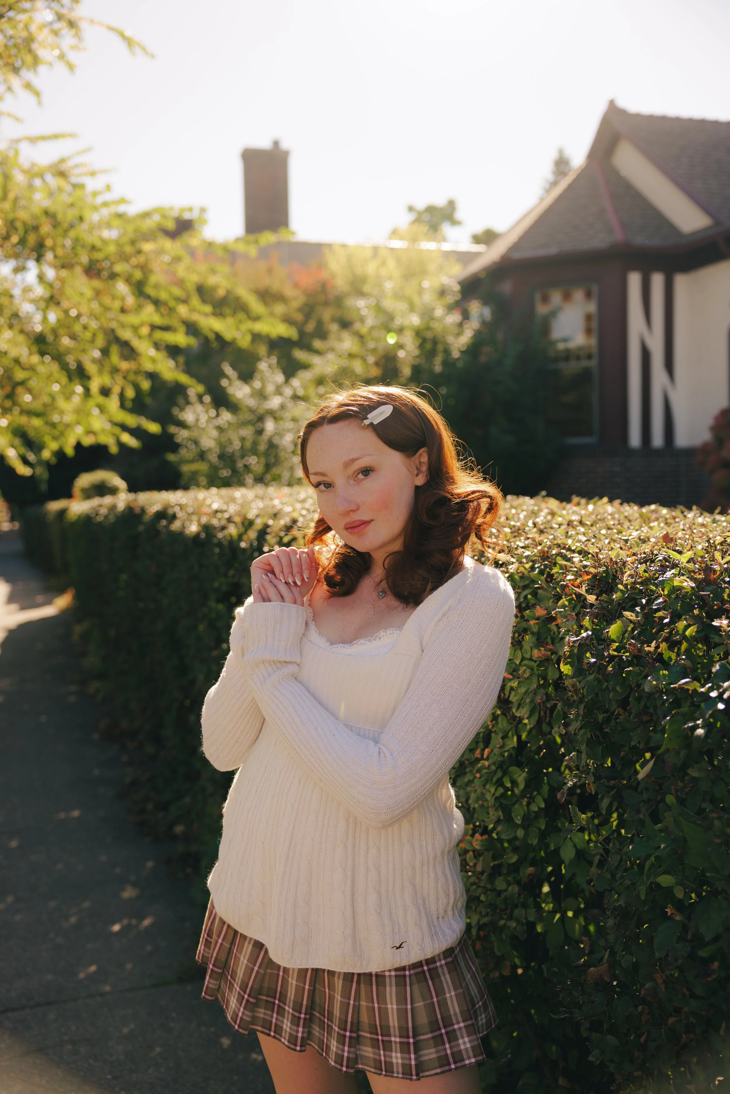 A young woman with shoulder-length brown hair wearing a cream sweater, plaid skirt, and a white hair clip posing outdoors next to a hedge, with houses and trees in the background on a sunny day.