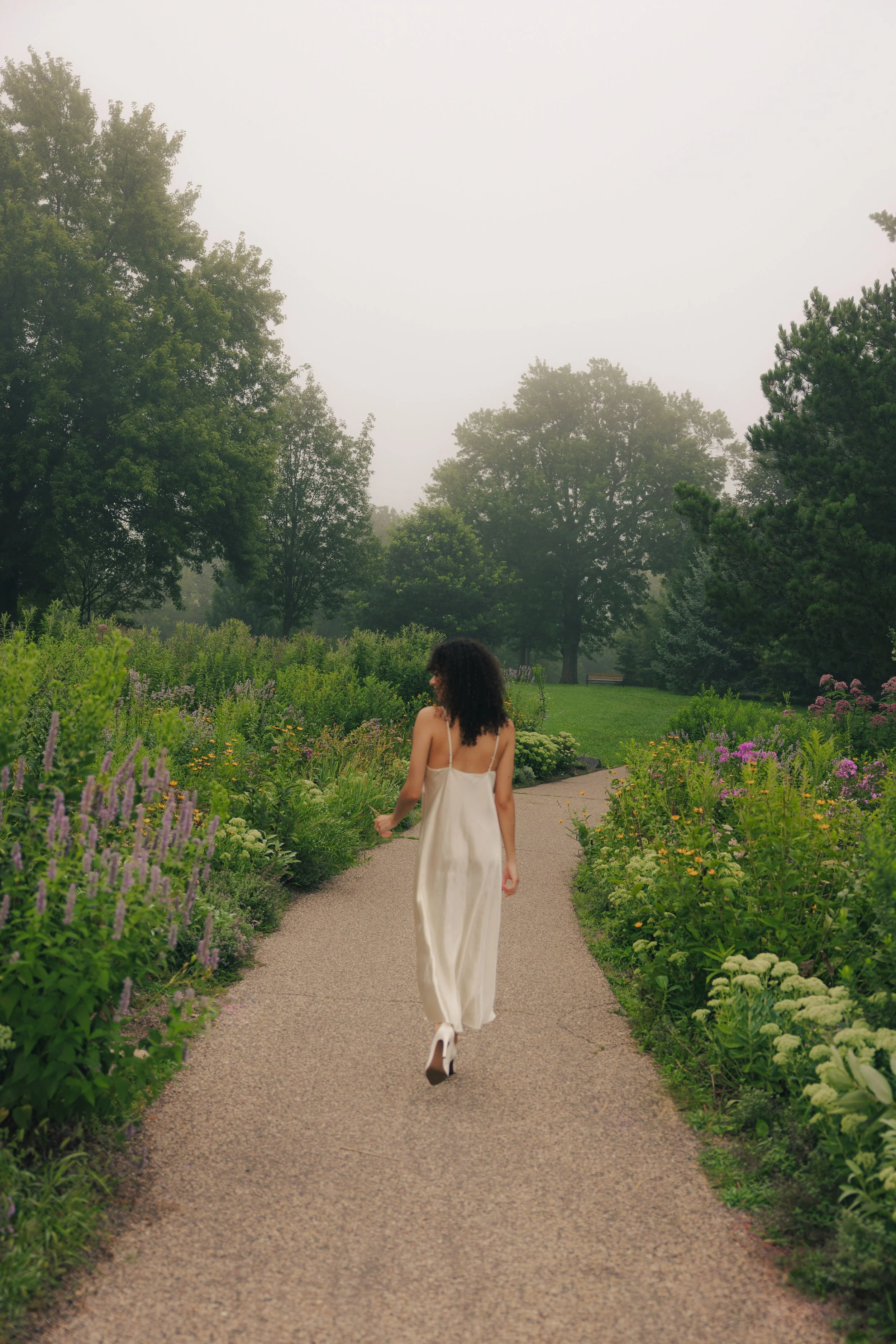 A woman in a white dress walking on a garden path surrounded by flowering plants.