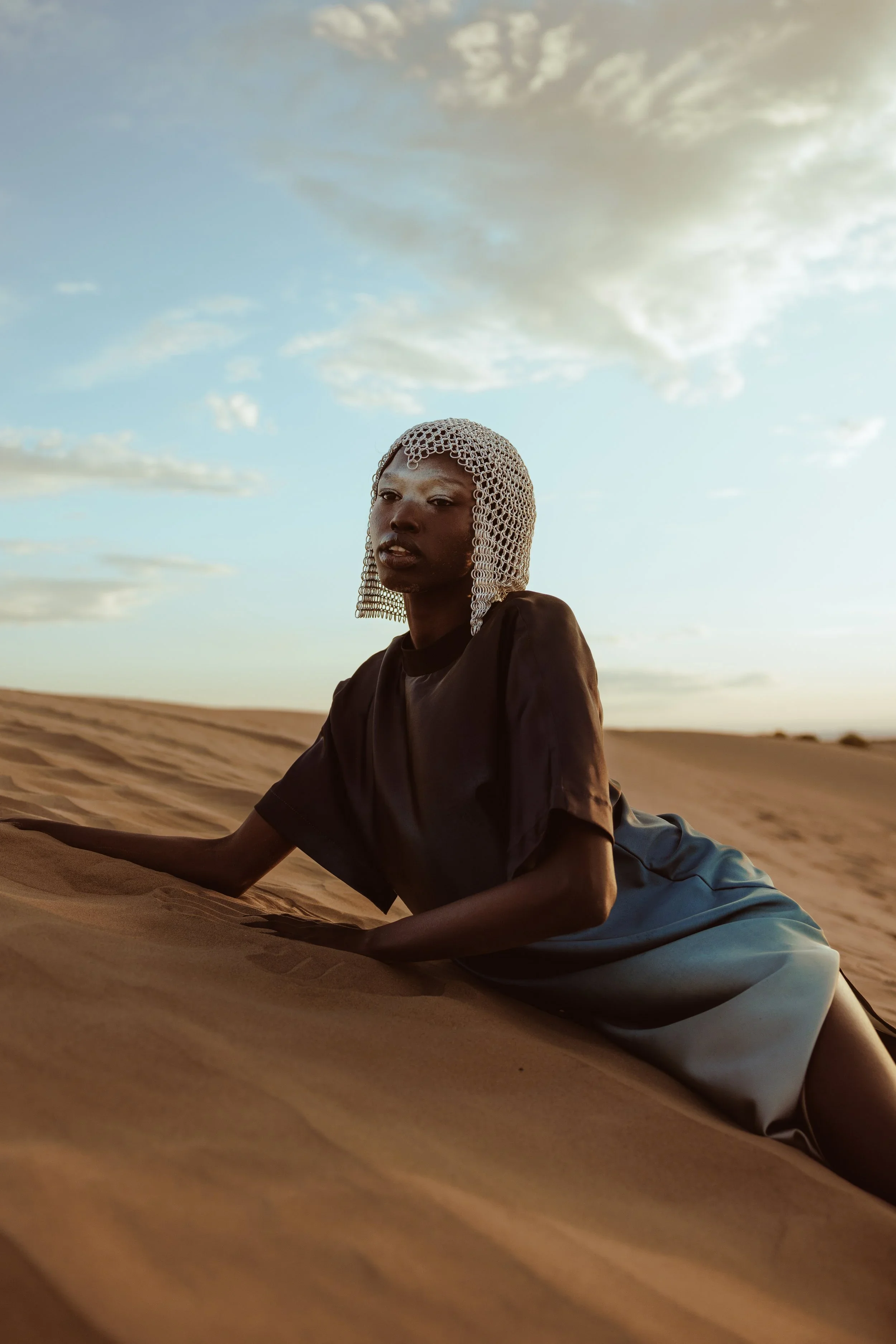 A woman with dark skin wearing a crochet helmet and dark clothing reclines on sand dunes in a desert landscape under a partly cloudy sky during sunset.