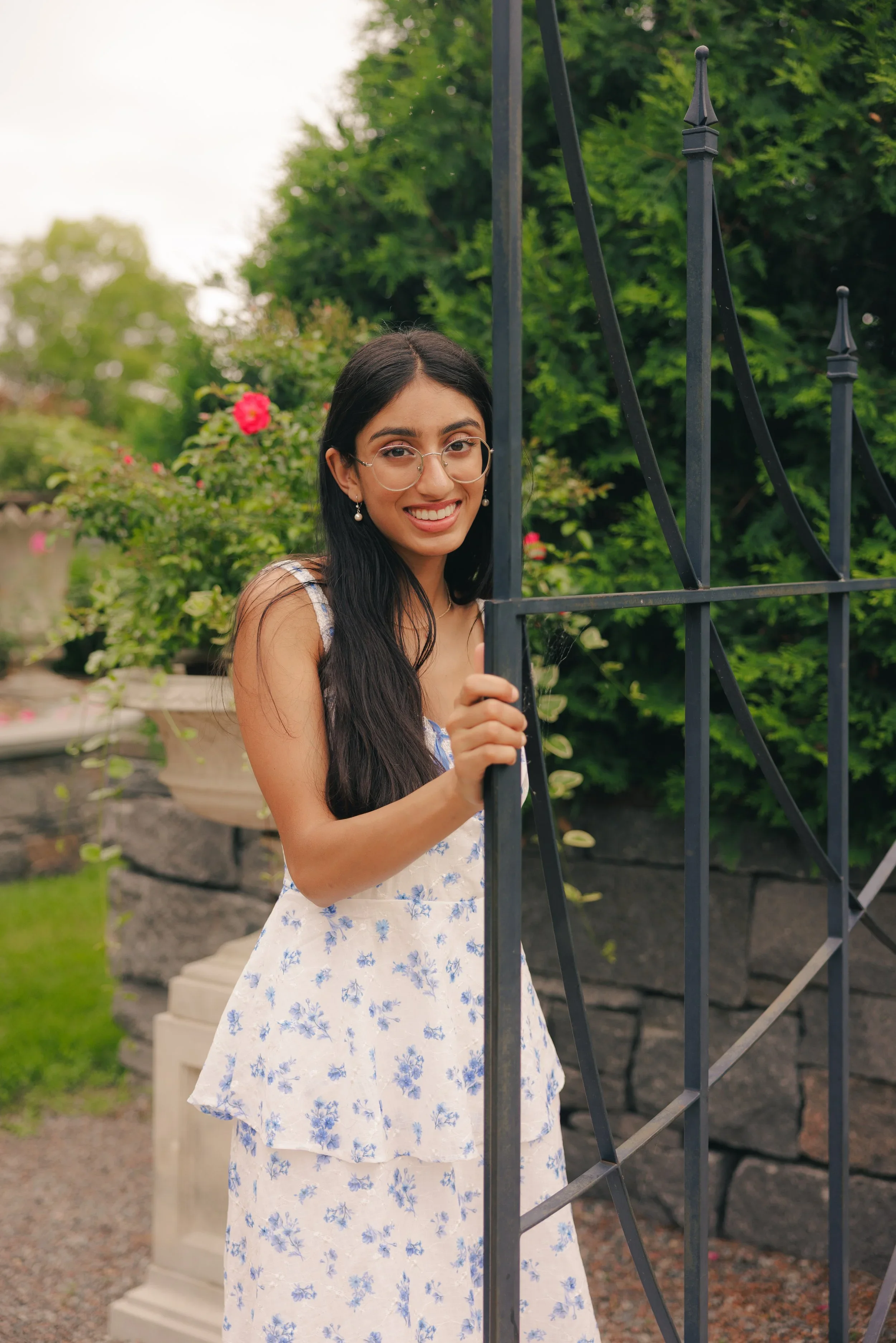 A young woman with long dark hair, wearing glasses and a floral dress, smiling while holding a black metal gate outdoors with green bushes and pink flowers in the background.