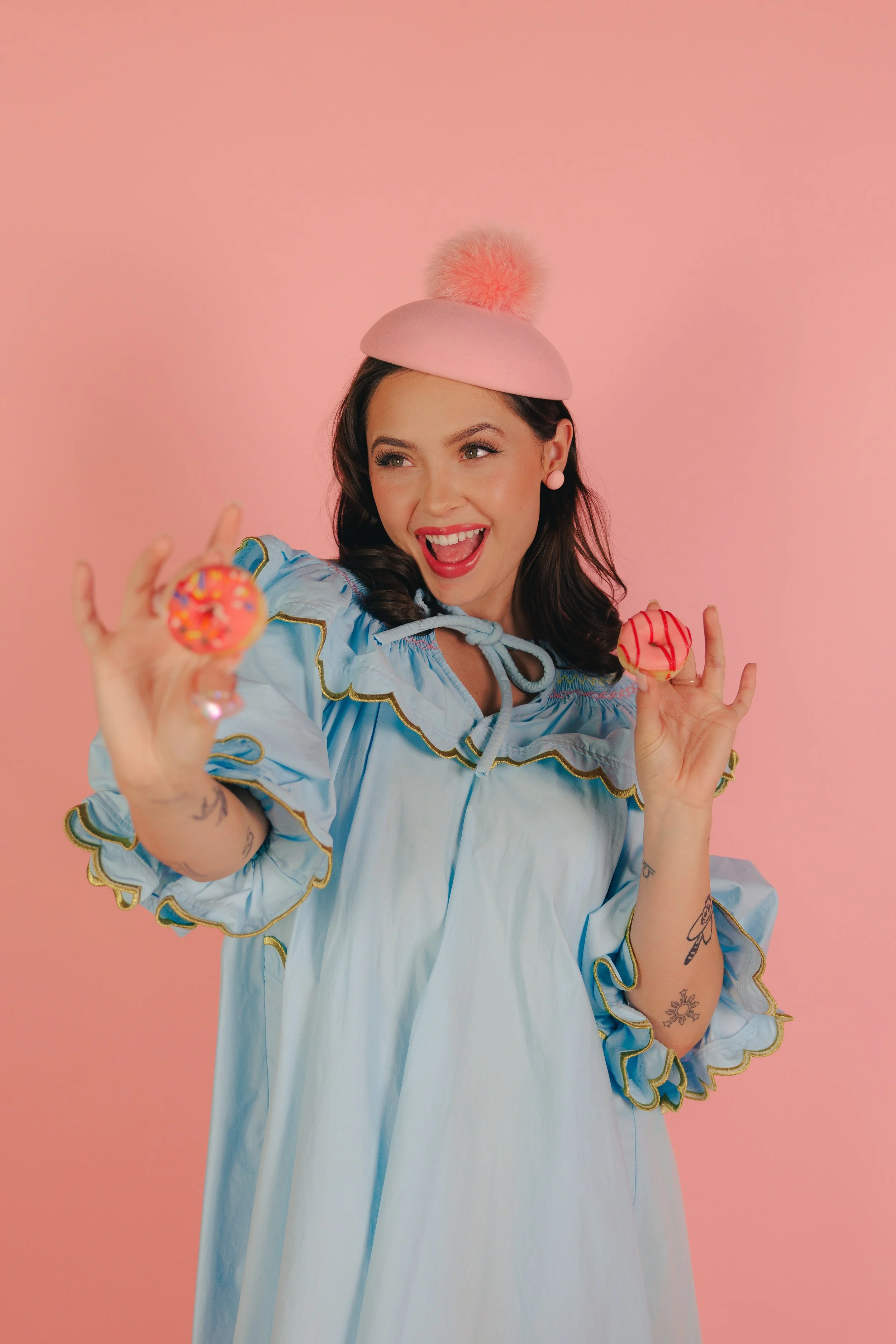 A woman with dark brown hair wearing a blue dress and pink hat holding two mini donuts