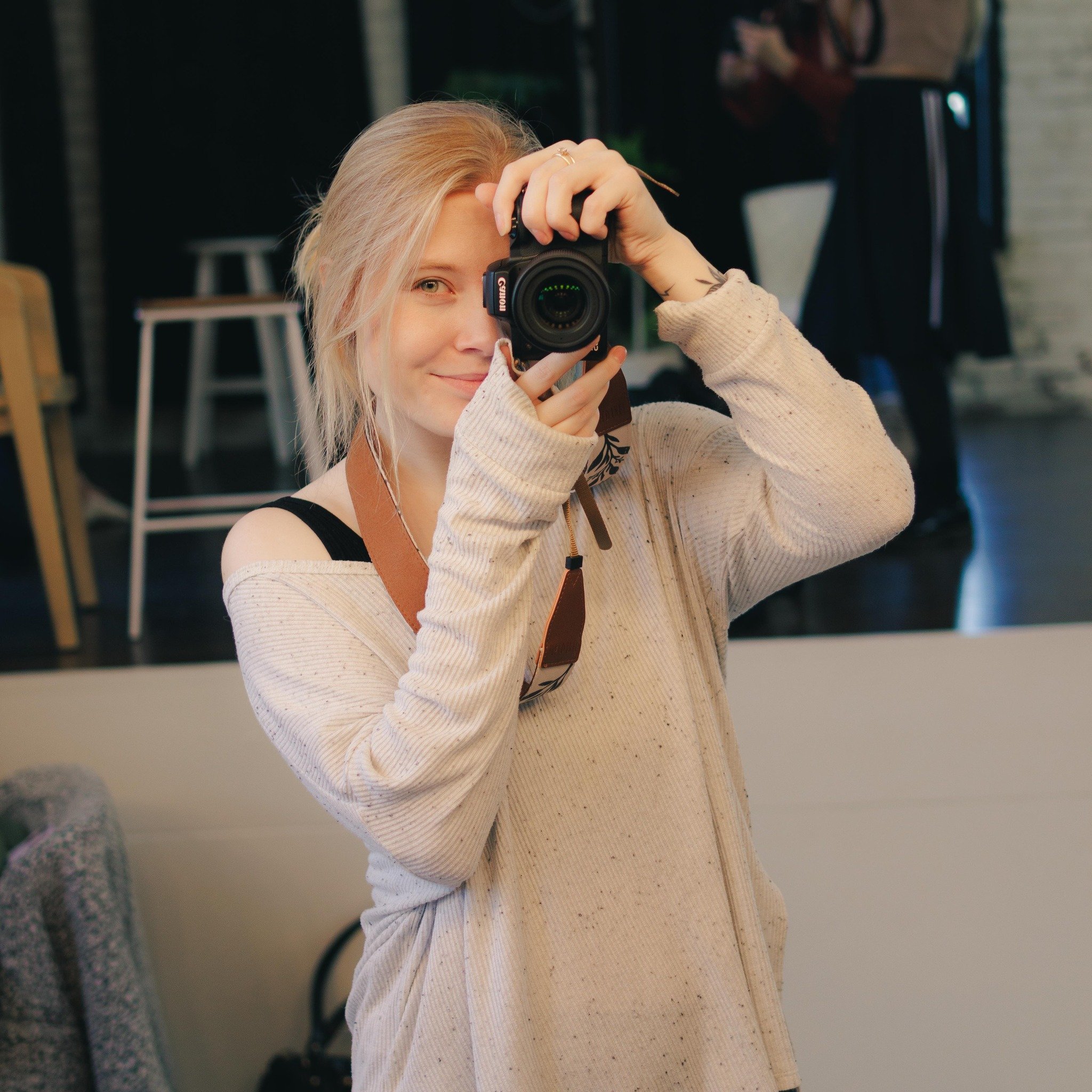 A young woman with blond hair and a light-colored sweater is taking a photo with a camera, looking through the viewfinder, in an indoor setting.