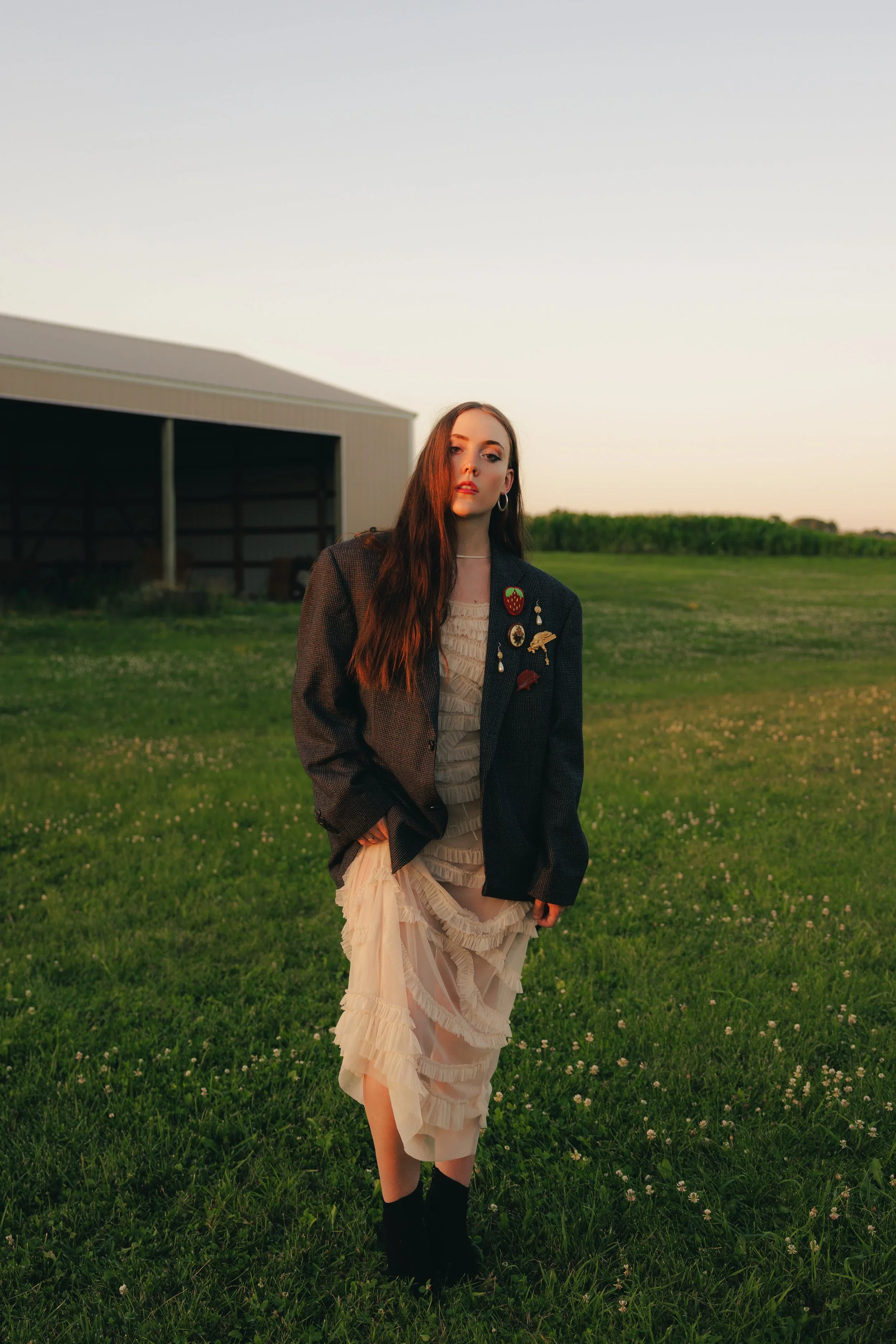 A woman with long brown hair and hoop earrings stands on a grassy field at sunset, wearing a layered beige dress, black boots, and a dark blazer adorned with pins.