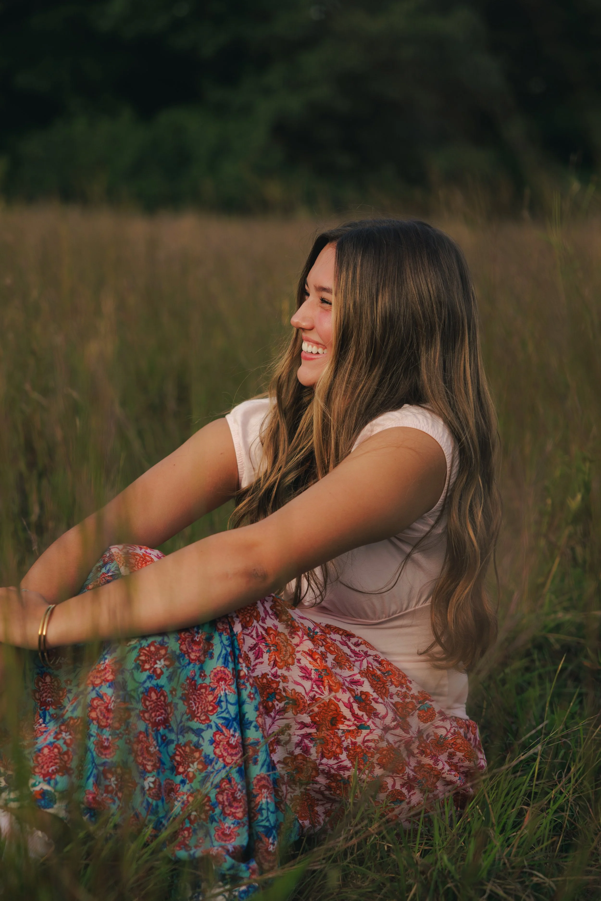 A young woman sitting in a grassy field during sunset, smiling and looking to the side, wearing a light pink shirt and a colorful, patterned skirt.