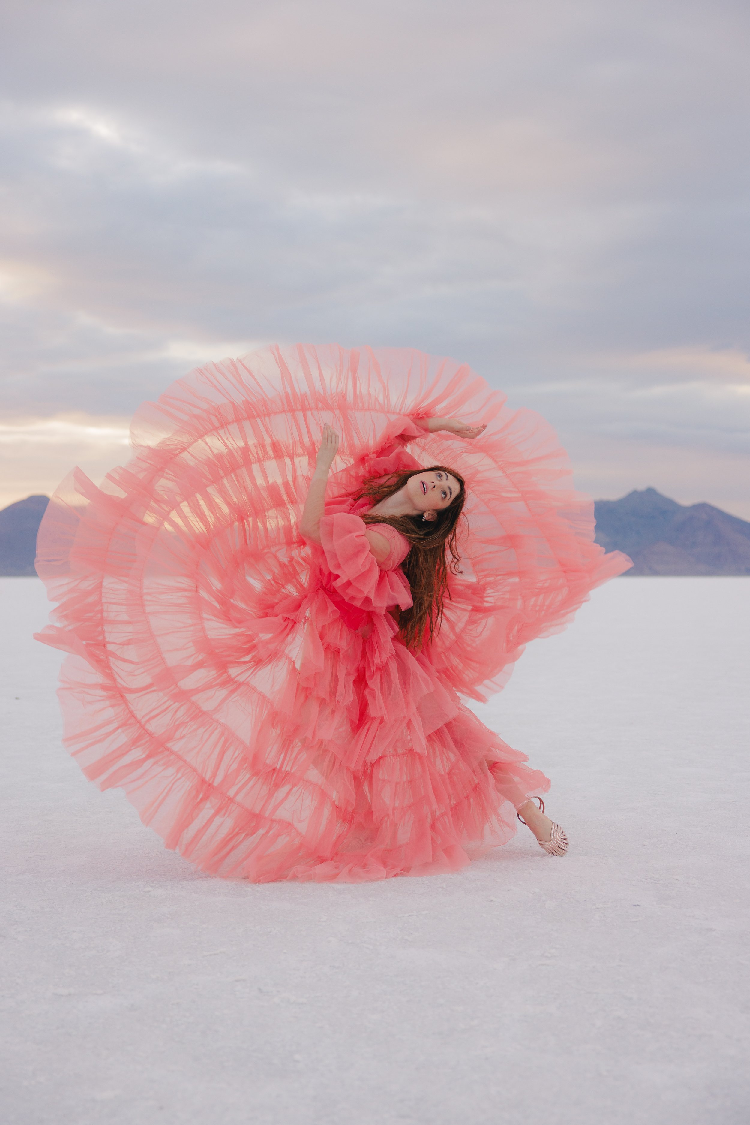 A woman dancing outdoors on a salt flat, wearing a flowing, ruffled pink dress with a large circular ruffle behind her, against a backdrop of mountains and a cloudy sky.