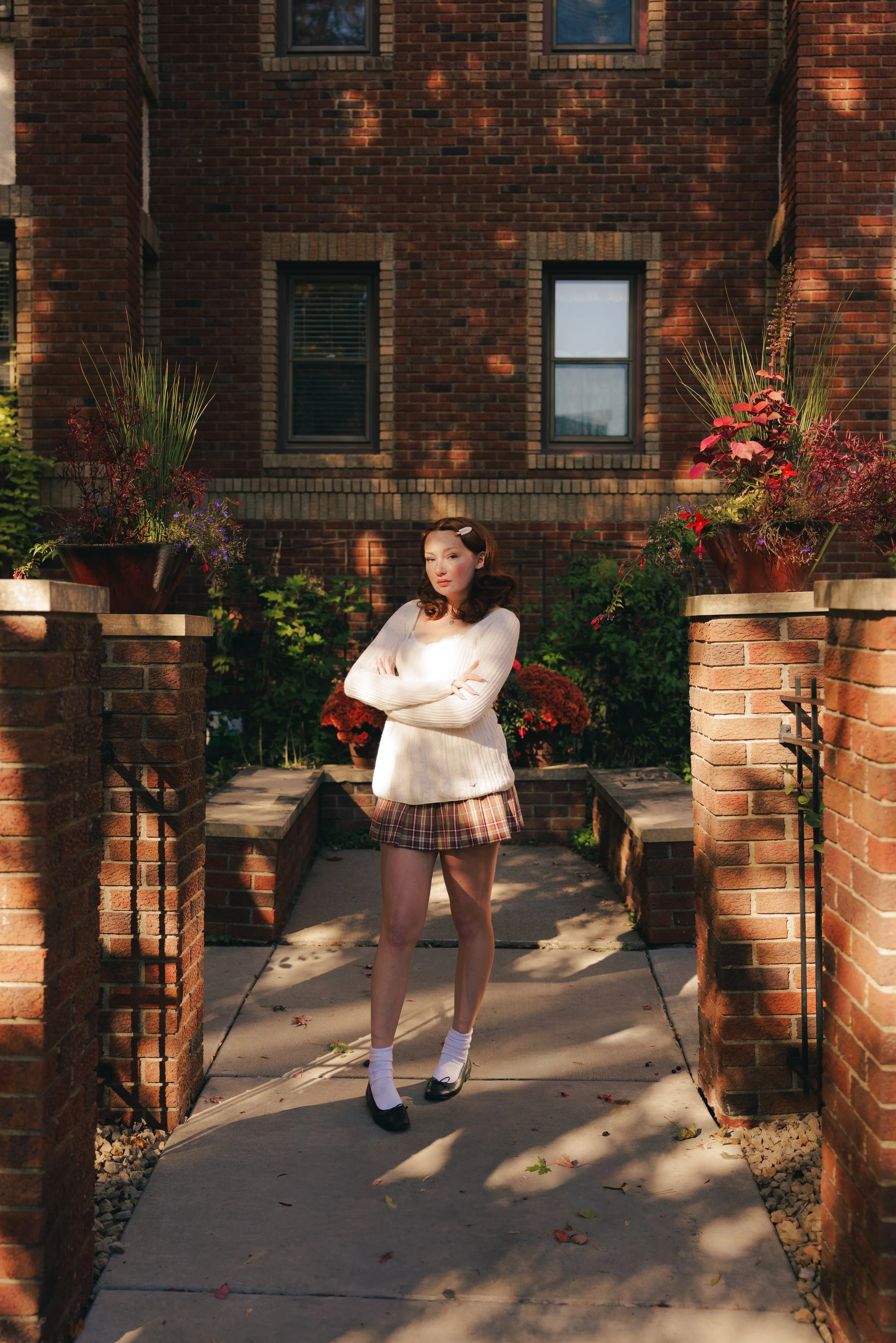 A woman standing with crossed arms in a garden entryway with brick walls, potted plants, and flowering bushes, sunlight casting shadows.
