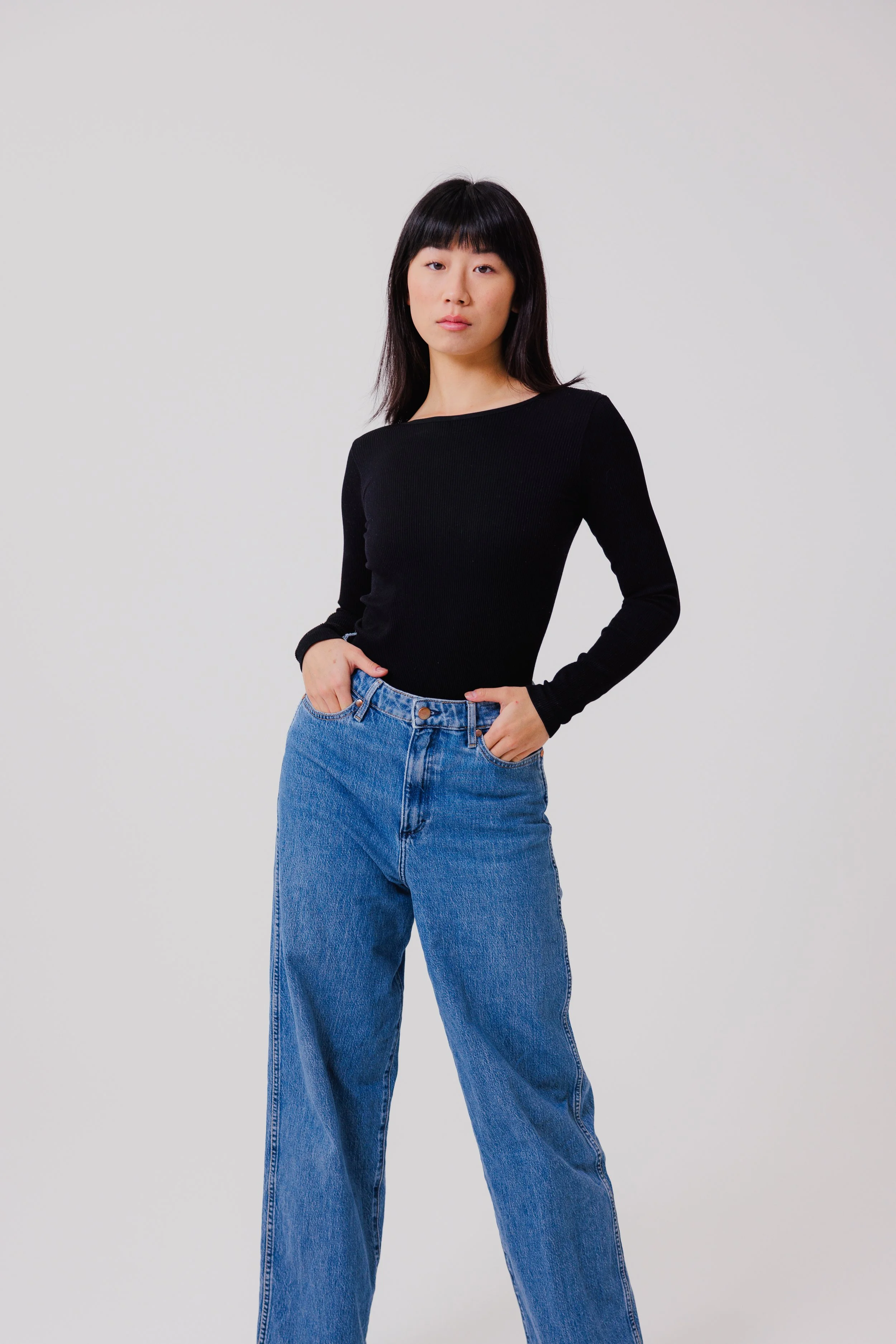 Young woman with black hair wearing a black long-sleeve top and blue jeans standing against a plain white background.