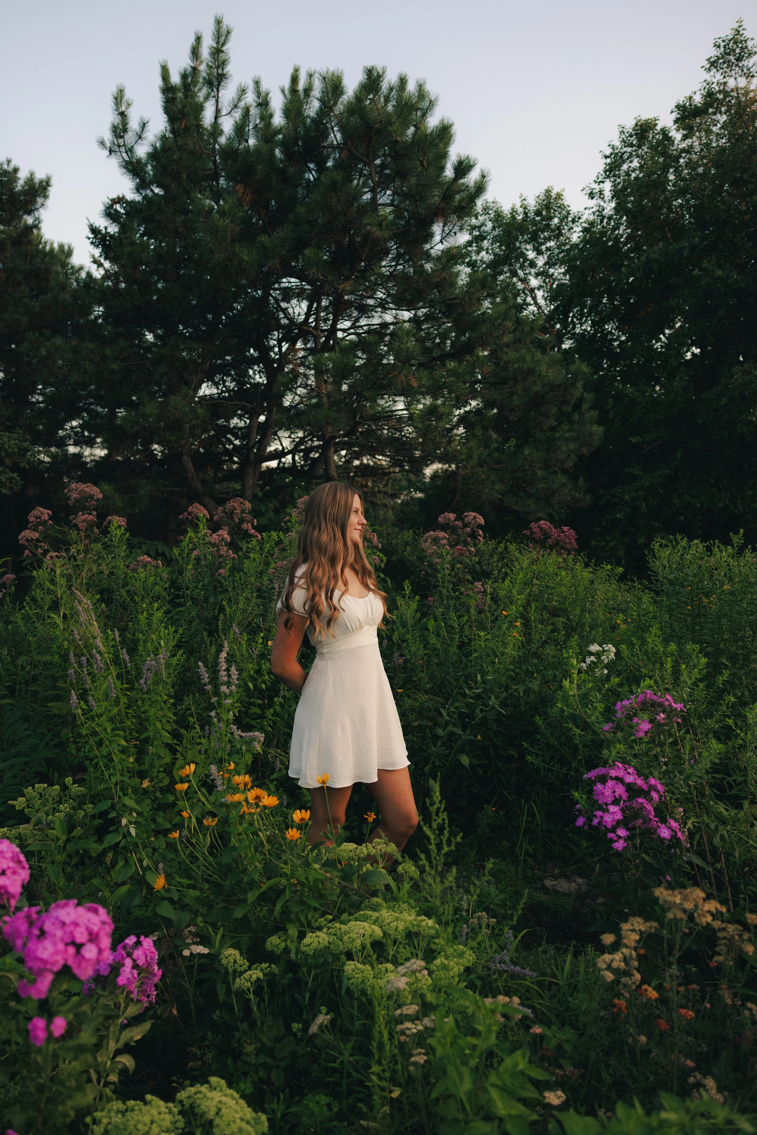 A young woman with long, wavy brown hair wearing a white dress stands among lush green plants and colorful flowers, with a large pine tree in the background.