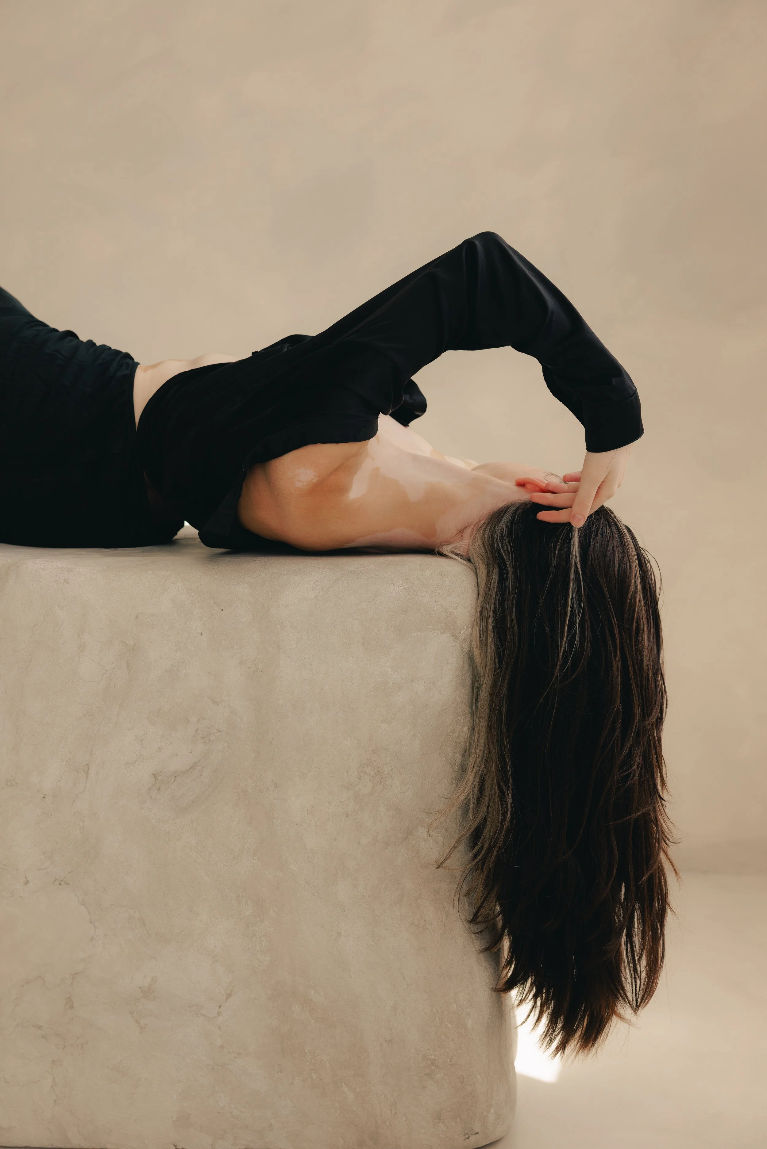 A woman with long brown hair is lying on a beige surface, wearing a black top and pants, with her head hanging upside down over the edge.