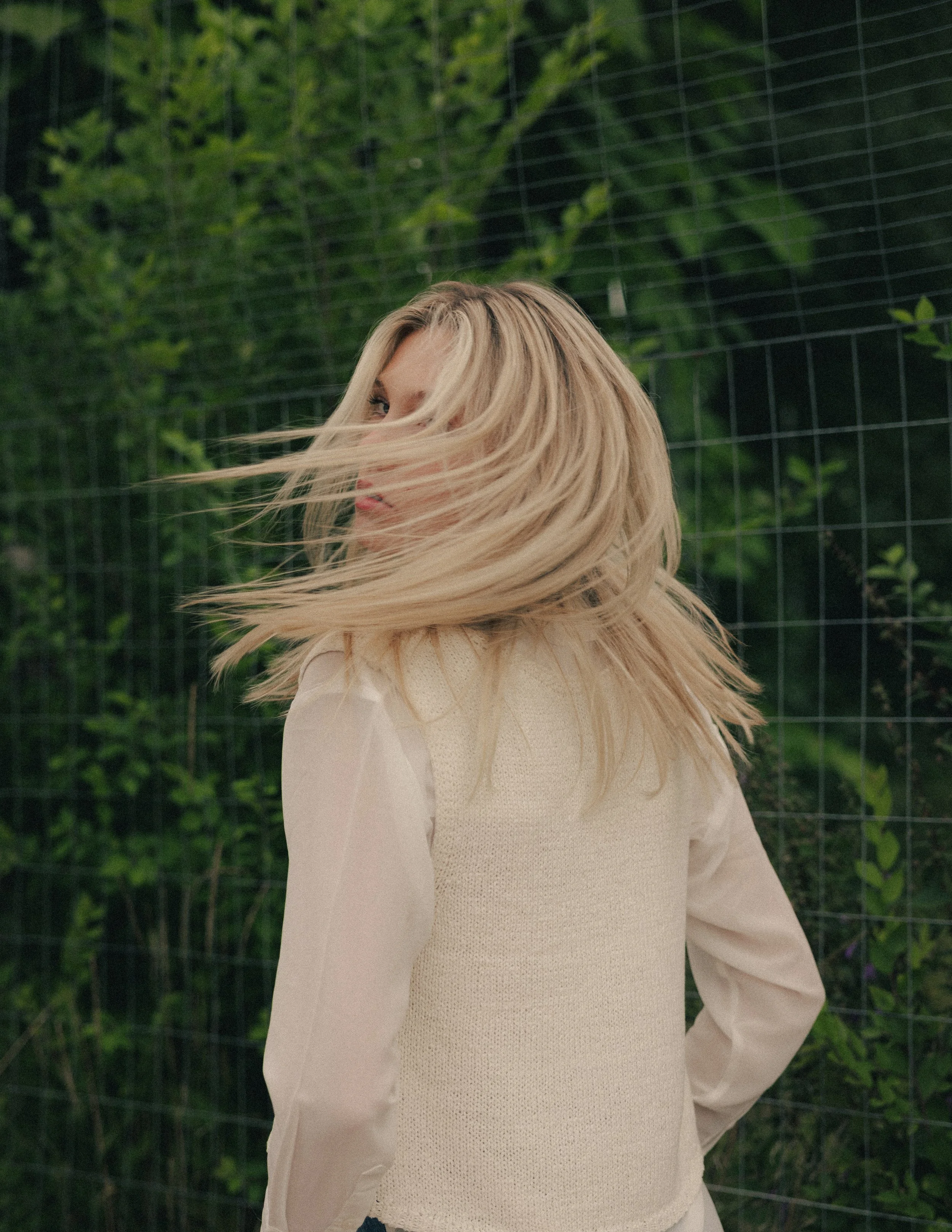A woman with long blonde hair wearing a beige sweater standing near a wire fence with green foliage behind her.