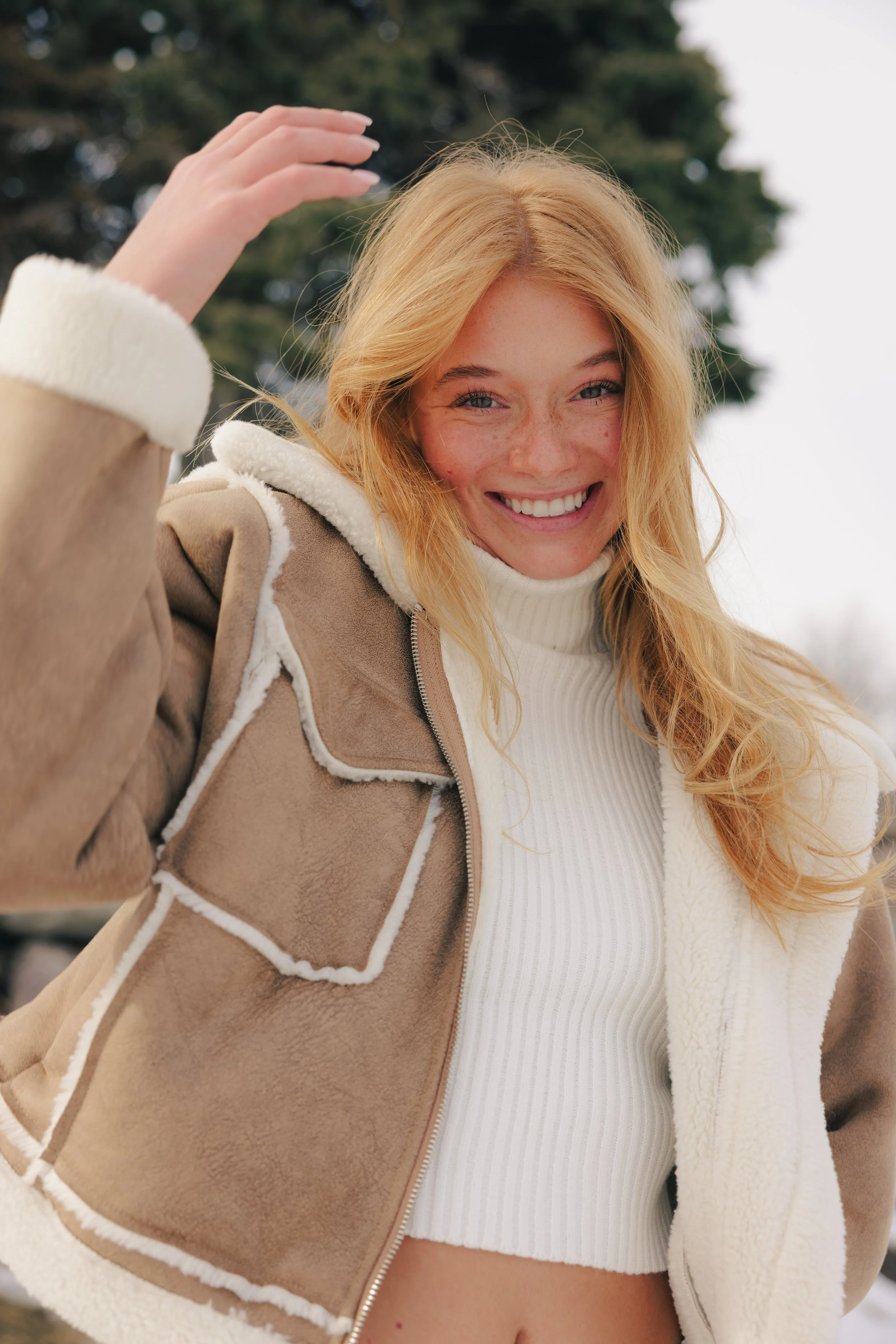 A young woman with long, wavy blonde hair smiling outdoors in a snowy environment, wearing a beige and white shearling jacket and a white turtleneck.