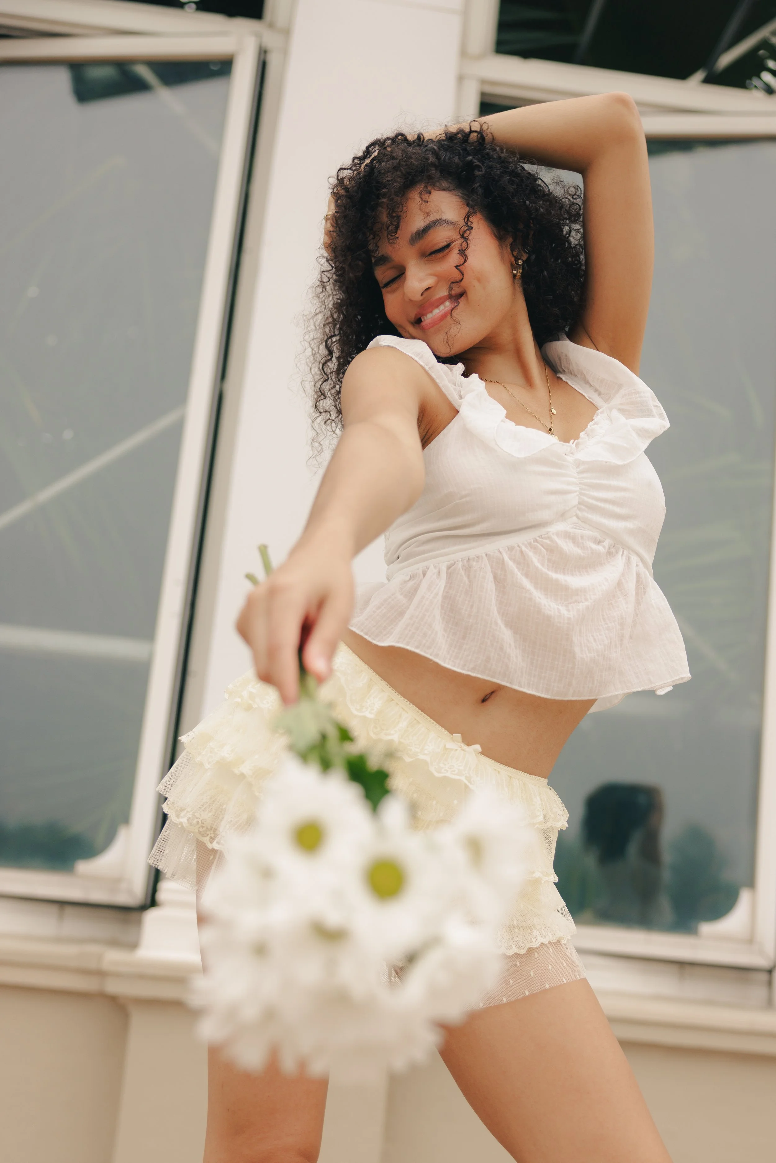 A woman with curly hair smiling and holding a bouquet of white daisies, wearing a cream-colored ruffled top and matching ruffled skirt, standing in front of a window.