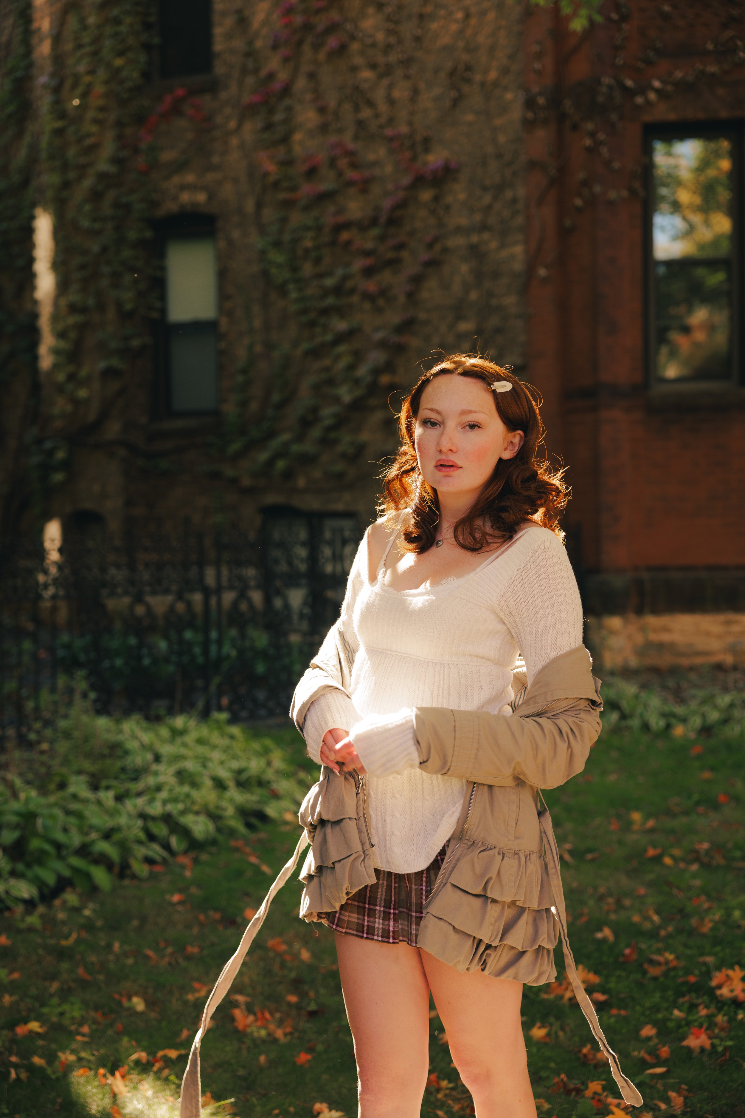 Young woman with red hair standing outdoors in front of a brick building covered in ivy, wearing a cream sweater, beige jacket, and plaid skirt.