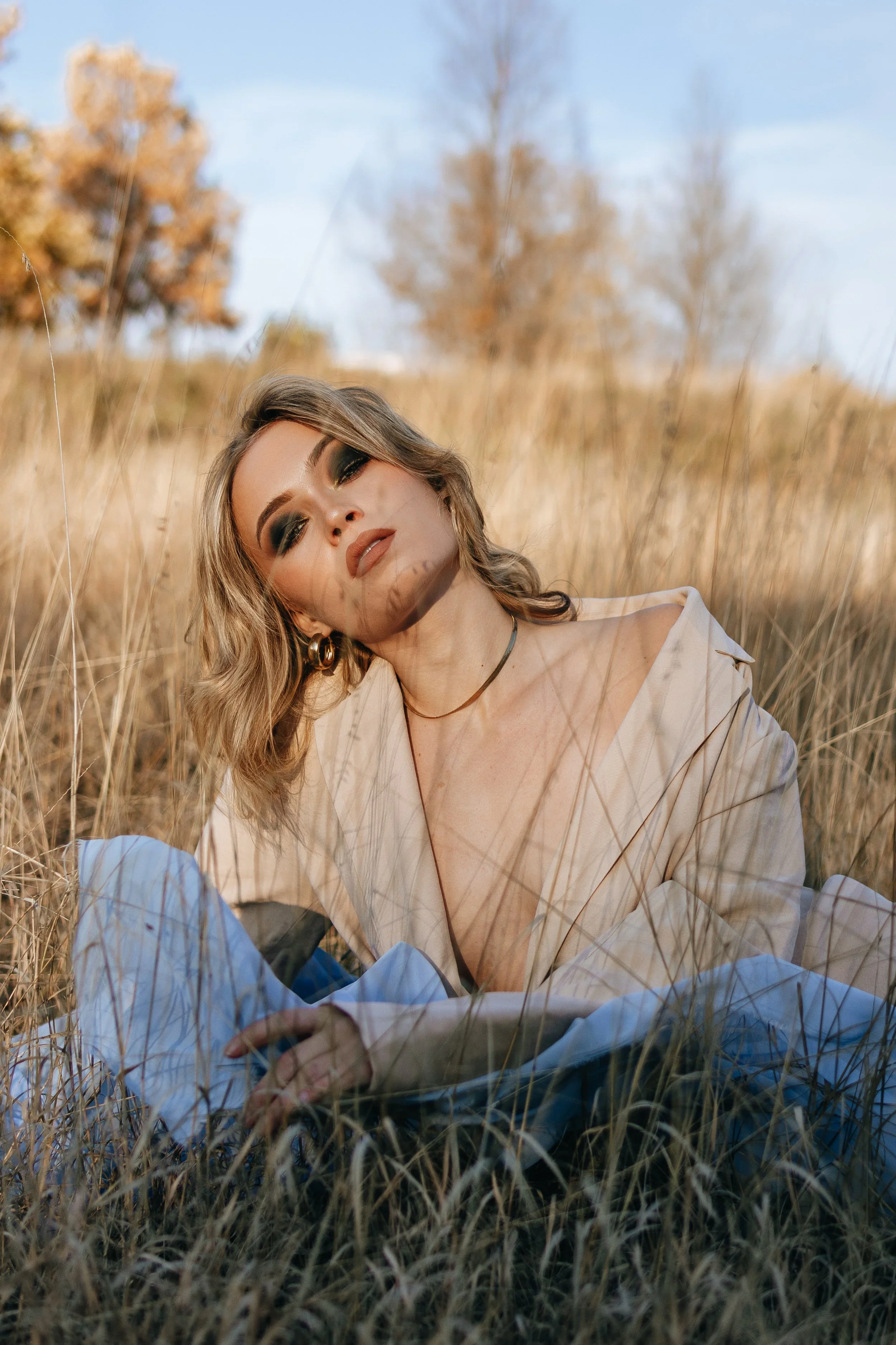 A woman with wavy blonde hair and makeup, wearing a beige coat, lying in tall grass in a field with trees and a blue sky in the background.