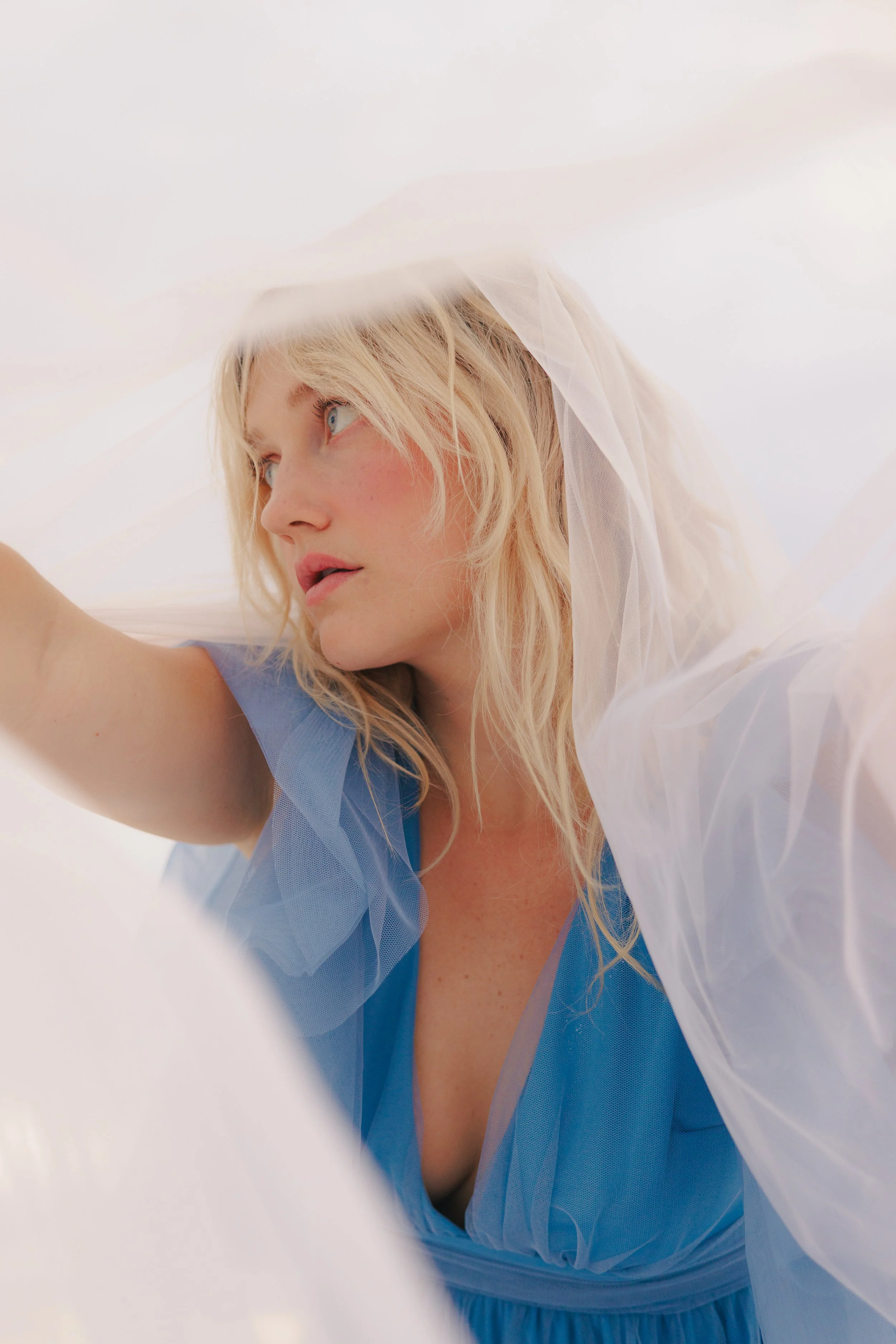 A woman with blonde hair and blue eyes wearing a blue dress, surrounded by sheer white fabric, looking to the side.