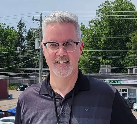 A middle-aged man with gray hair and beard, wearing glasses and a dark polo shirt, standing outdoors in front of trees and a parking lot.