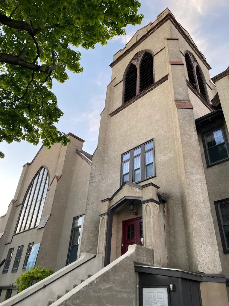 A tall beige building with gothic-style arched windows and a pointed roof, partially covered by green leafy trees, with a red door at the top of stairs leading up to the entrance.