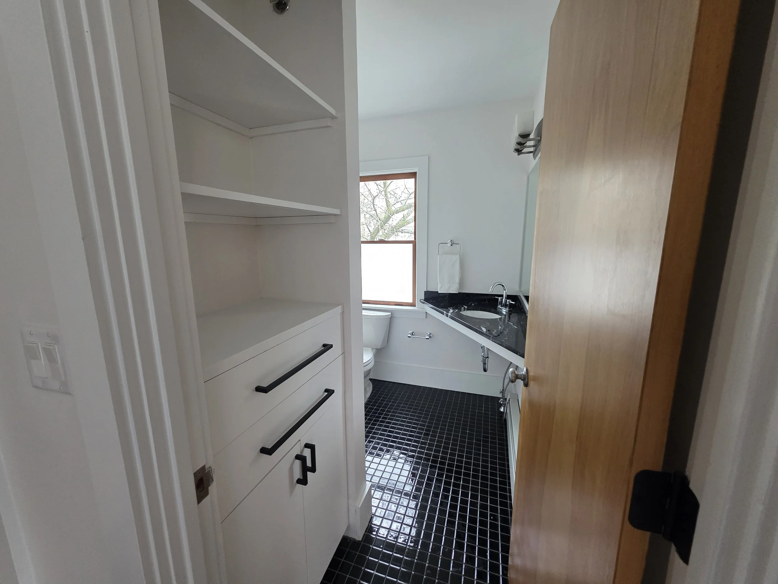 View of a modern bathroom with white shelves, a window showing tree branches, and a wooden door. There is a black sink and a toilet visible, with black tile flooring.