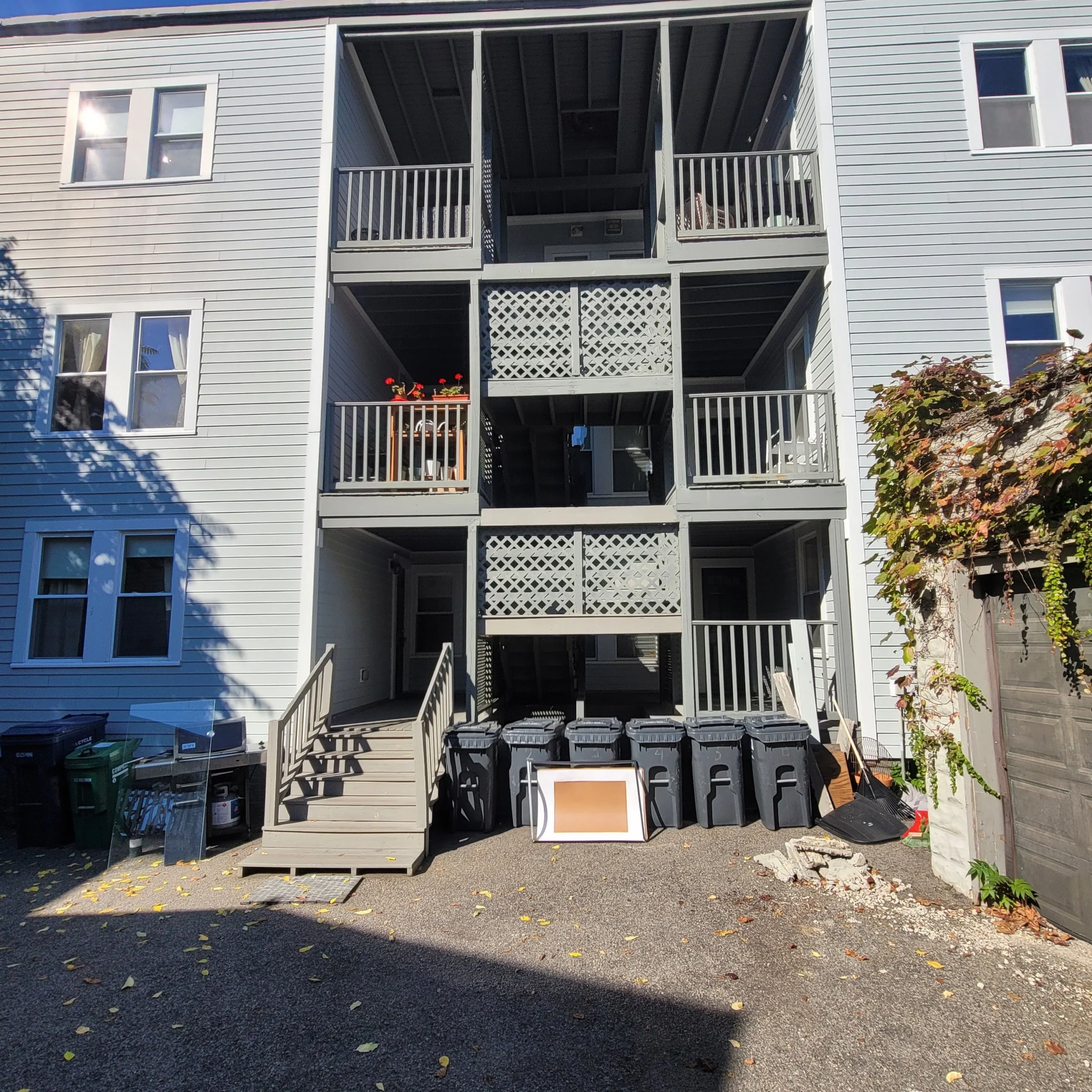 A multi-story residential building with white siding and multiple balconies. Some balconies have flower pots and furniture. The ground is covered in fallen leaves, and there are garbage bins at the bottom. A tree with autumn leaves is on the right side of the image.