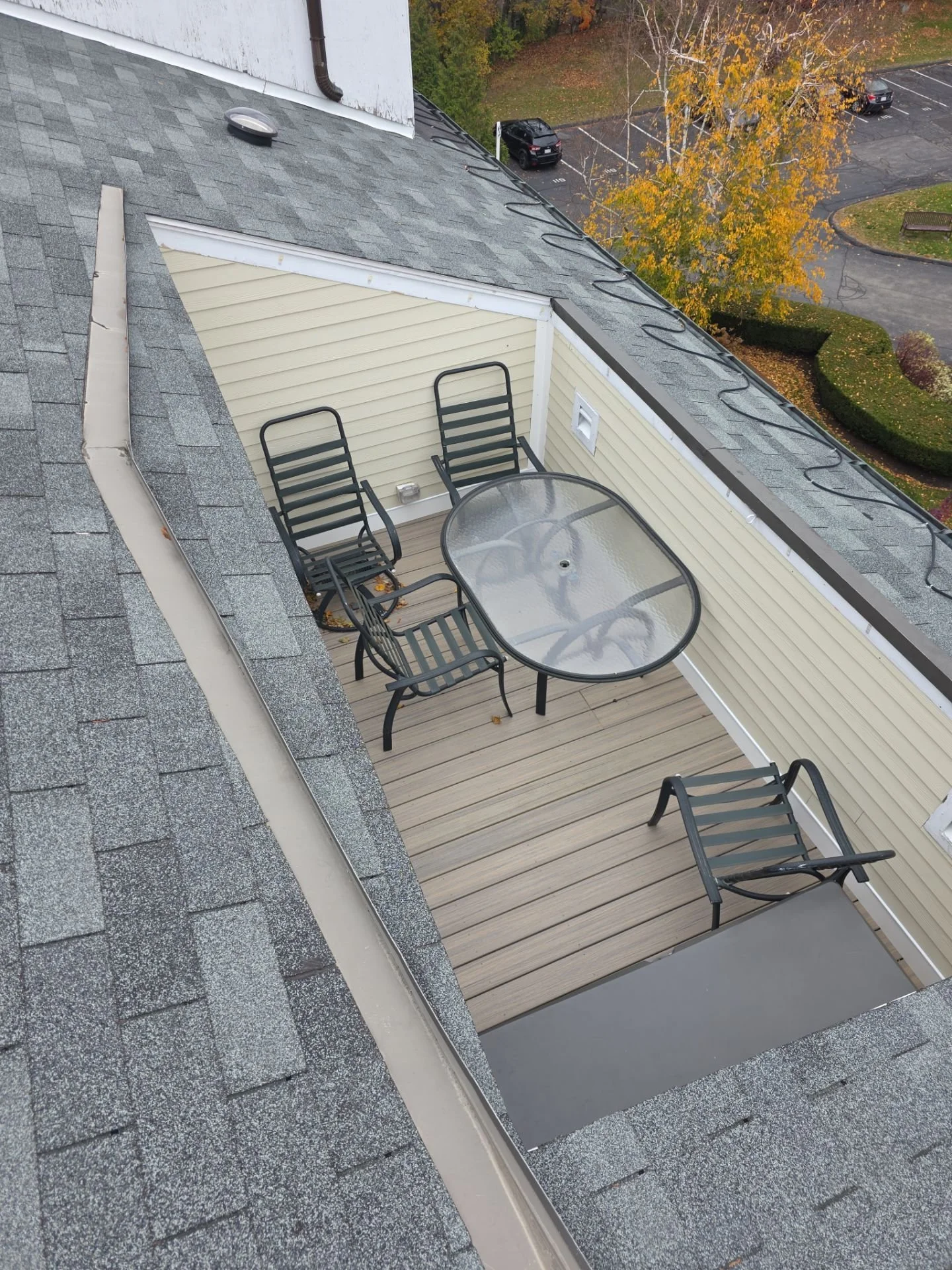A rooftop view showing a small balcony with four black chairs and a glass table, beige siding, gray shingles on the roof, and a parking lot with two cars and a tree with yellow leaves in the background.