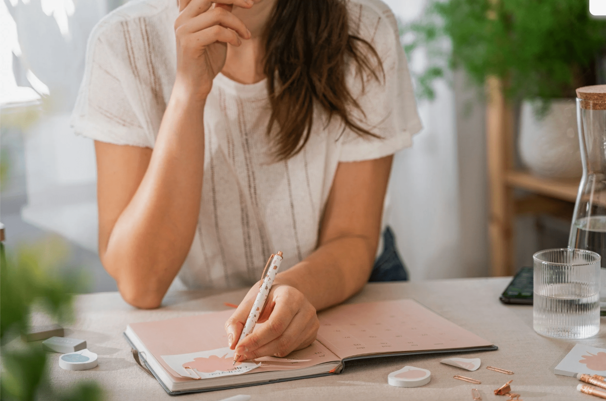 A woman with brown hair wearing a white t-shirt is sitting at a desk, writing in a pink planner with a polka dot pen. On the desk are decorative pins, a glass of water, a jar of water, and a smartphone. The background shows a plant and bookshelves.