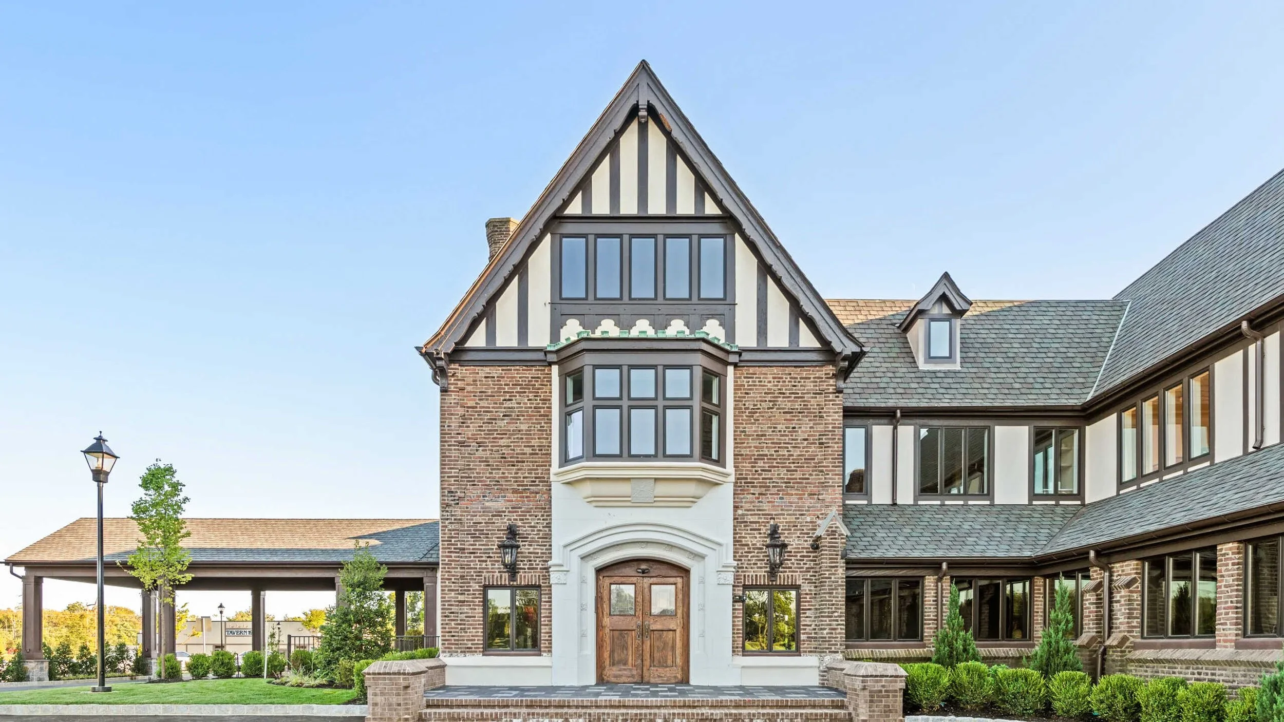 Exterior view of a grand Tudor-style golf club: red brick, white stucco with dark timber framing, and a gabled roof. A stone-arched wooden entryway serves as the focal point, flanked by manicured green shrubs and a covered driveway to the left.