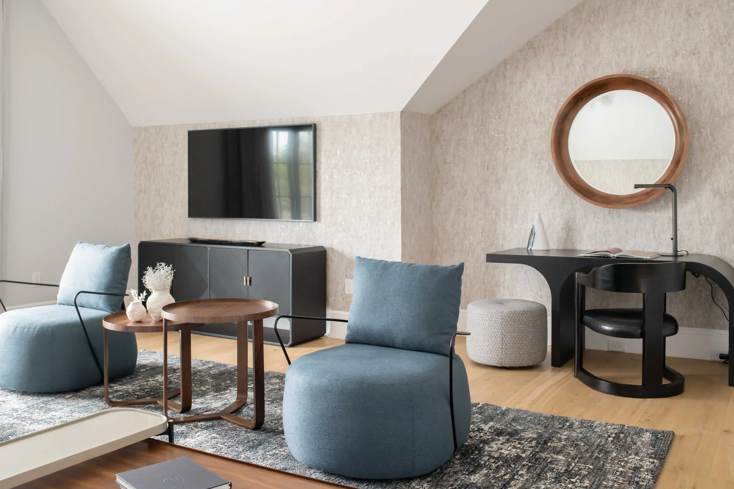 Contemporary sitting area with dusty blue modular seating, round wood side tables with black legs, black sculptural console, and textured neutral wallcovering under sloped ceiling with black-framed doors.
