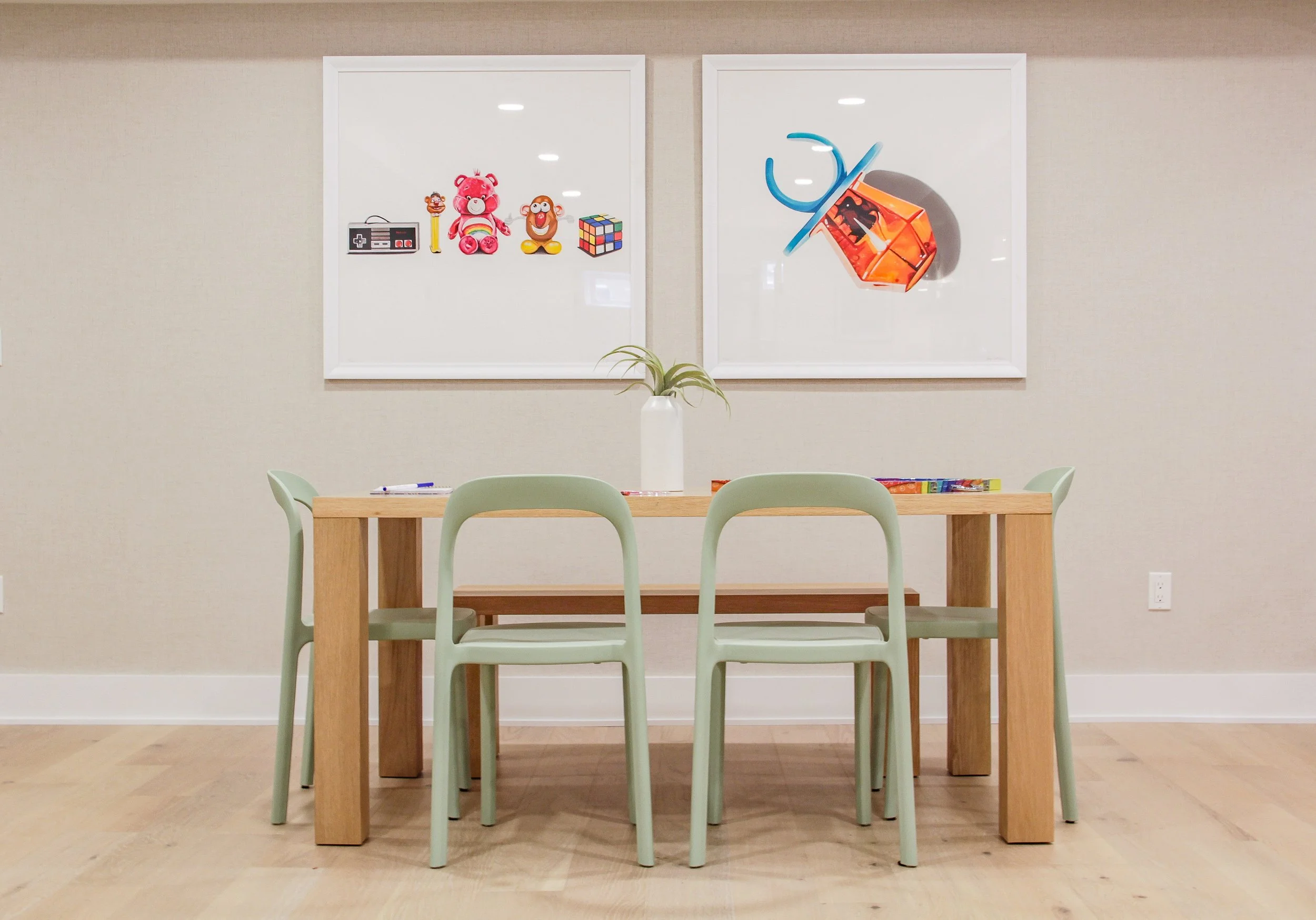 Children's activity area with natural wood table surrounded by sage green molded plastic chairs, pair of framed prints above showing vintage toys including Care Bear and Rubik's Cube, and orange View-Master toy, with small air plant in white vase as 