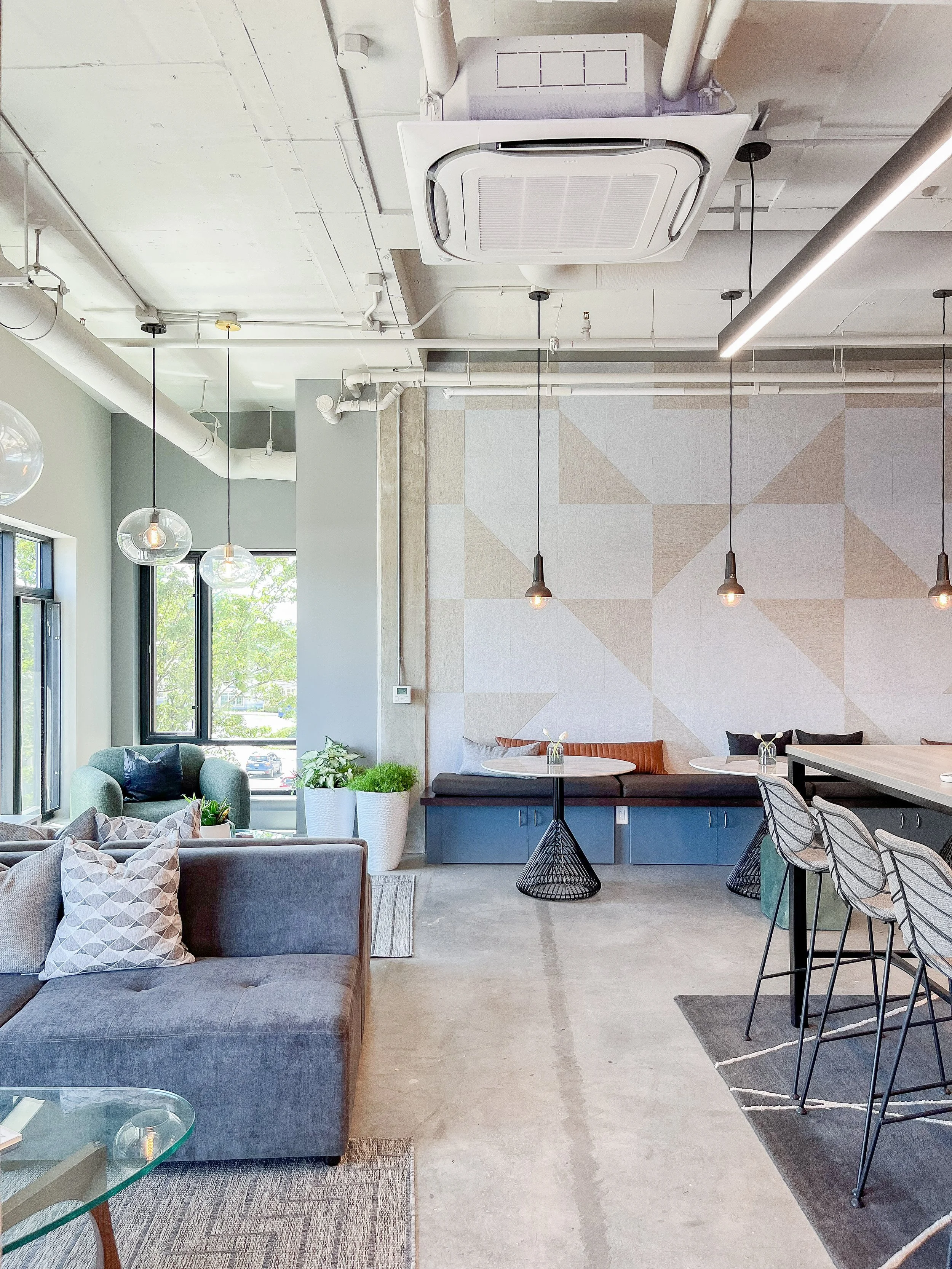 Collaborative workspace with two navy blue high-top tables surrounded by wire-frame counter stools on gray geometric area rug, large-scale triangular acoustic wall panels, wood vertical slat divider, wall-mounted TV flanked by blue framed artworks, m