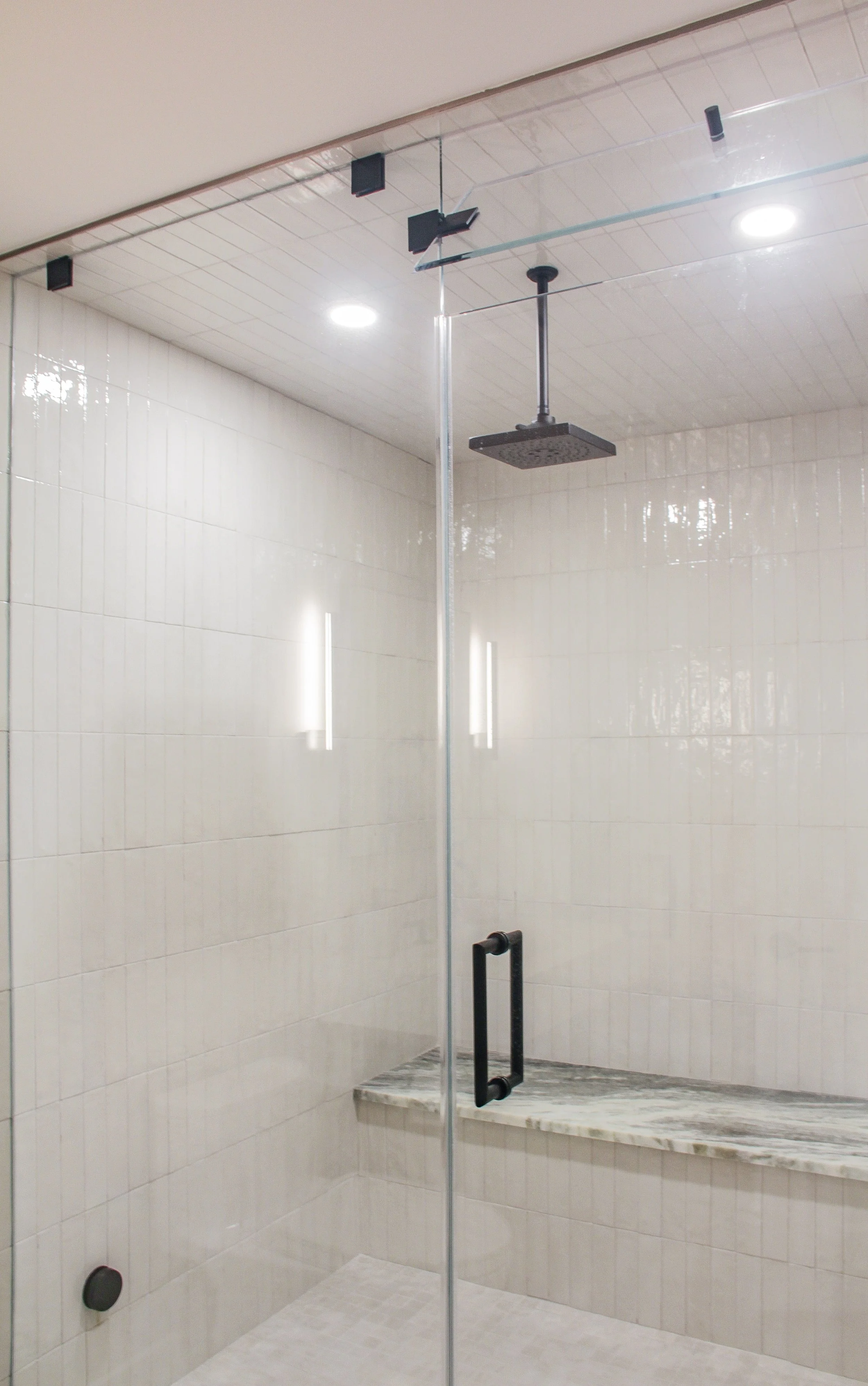 Walk-in shower with floor-to-ceiling white subway tile in vertical layout, frameless glass door with matte black hardware, ceiling-mounted black rainfall showerhead, built-in marble bench, and white tiled ceiling.